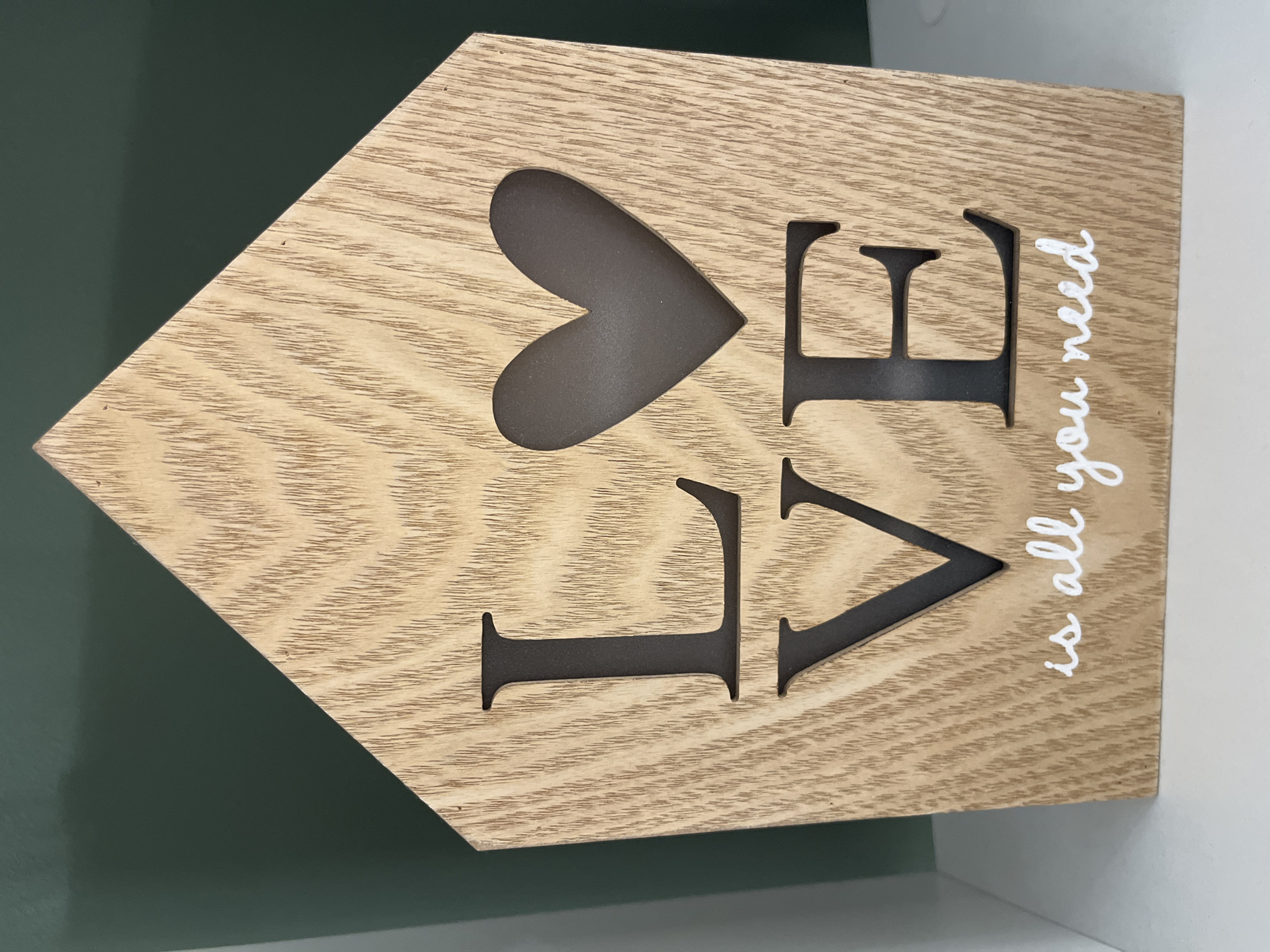 A wooden house-shaped decorative item with the word "LOVE" cut out of it is displayed on a white shelf against a dark green background. The word "LOVE" is spelled out with the letters L, V, and E in black, with a heart shape cut out in place of the O. Below the word "LOVE," the phrase "is all you need" is written in white cursive text. The wooden surface has a prominent grain pattern. The item is positioned in a way that it is slightly angled towards the viewer. The scene does not provide any information to determine its location in Kharkiv, Ukraine.