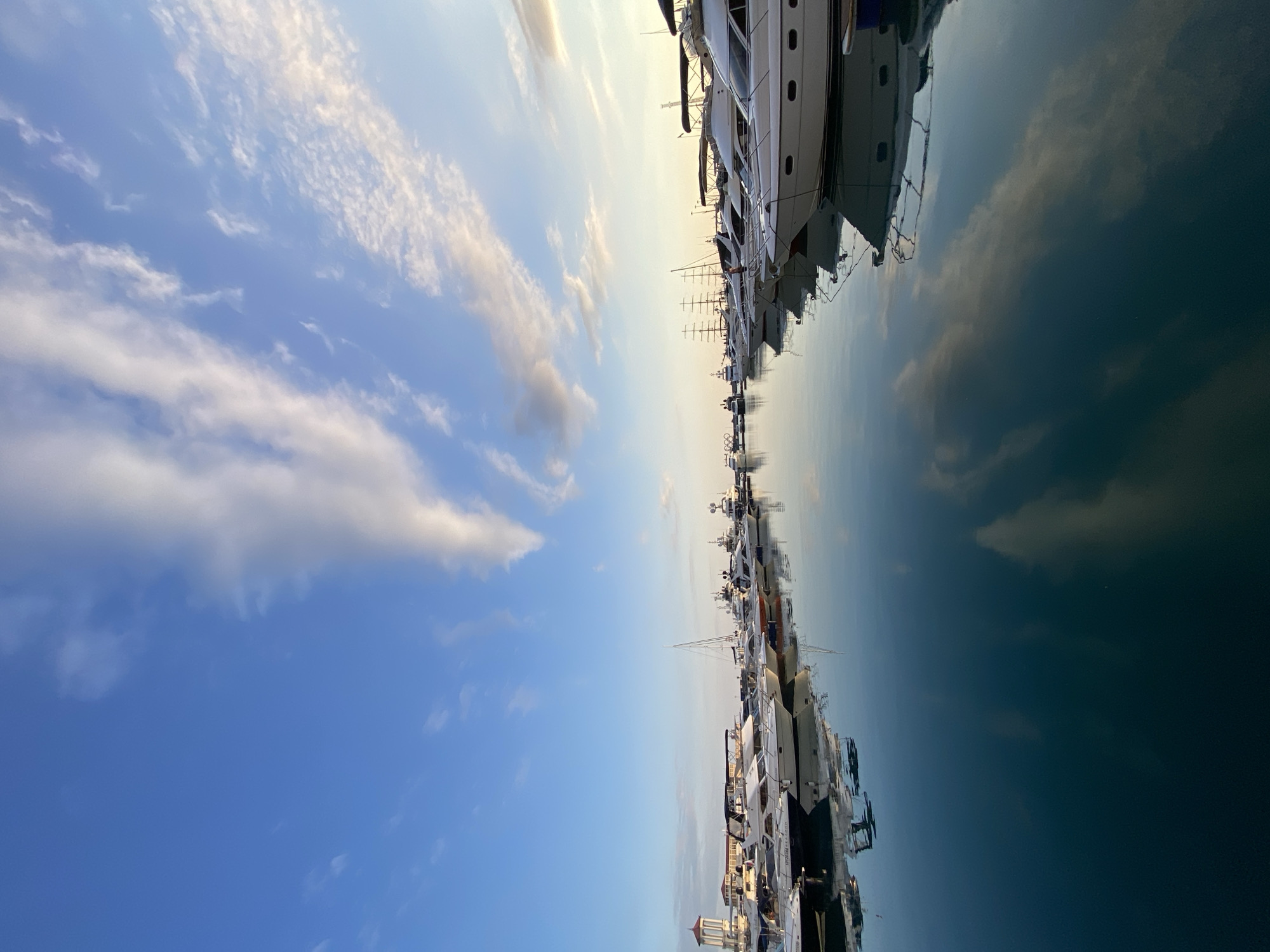 The image depicts a serene marina scene in Sochi, Russia, at what appears to be either sunrise or sunset, given the soft lighting and the angle of the sun. The sky is a clear blue, adorned with scattered, wispy white clouds that are gently reflected in the still, tranquil water of the harbor.
A prominent feature of the scene is the multitude of yachts and boats docked neatly along the pier. These vessels, predominantly white and sleek, vary in size and design, suggesting a well-established harbor for leisure craft. Some of the yachts have dark roofs or covers, providing contrast against their white hulls. In the background, the outlines of more boats, including a tall-masted sailing ship, are visible, adding depth to the composition.
On the left side of the frame, a distinctive building with a domed roof and tower stands out, hinting at the architectural style of the area. The reflections of the boats and the sky in the water are nearly perfect, creating a symmetrical visual effect that enhances the calmness of the scene. The water is a deep teal, with subtle variations in shade reflecting the sky and the vessels. There are no people visible in the image, contributing to the peaceful and unpopulated atmosphere of the marina. The overall impression is one of order, luxury, and quietude, typical of a high-end yachting destination.