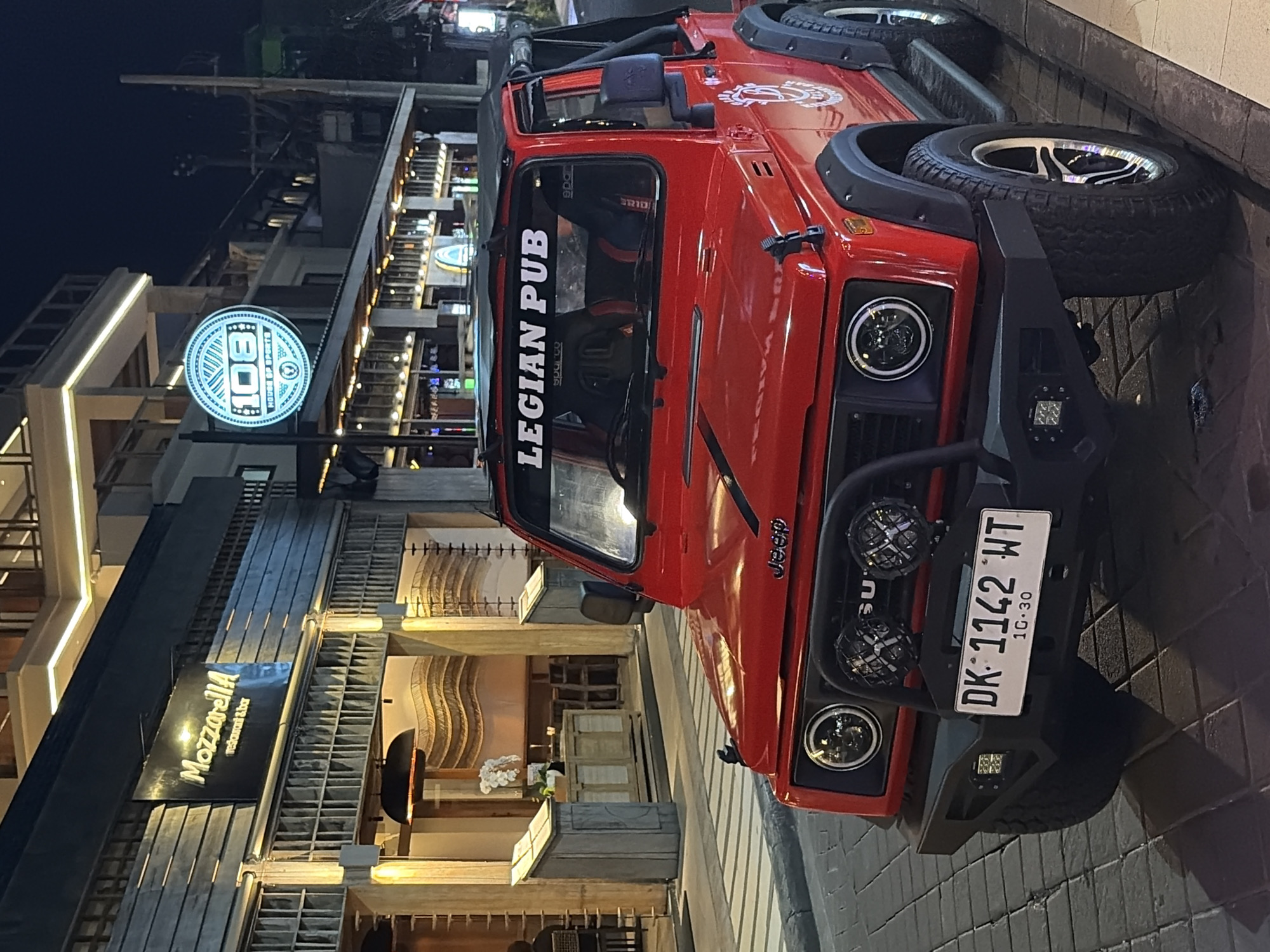 The image shows a red Suzuki Samurai parked on a cobblestone street at night in Kuta, Indonesia. The vehicle is facing the camera and is in the foreground. It has a black soft top, a black bull bar with auxiliary lights, and a license plate that reads "DK 1142 WT". A sticker on the windshield says "LEGIAN PUB".
Behind the car, a building with a modern facade is visible. A sign with "Mozzarella restaurant & bar" is illuminated on the left side of the building. To the right, a circular neon sign with "108 HOUSE OF SPORTS" is lit up. The building appears to be a commercial establishment with multiple levels, featuring balconies and architectural lighting. The street is paved with cobblestones, and there are streetlights and other ambient lighting illuminating the scene. The overall atmosphere is that of a lively street in a tourist area at night. No people are clearly visible in the immediate foreground or interacting with the vehicle.