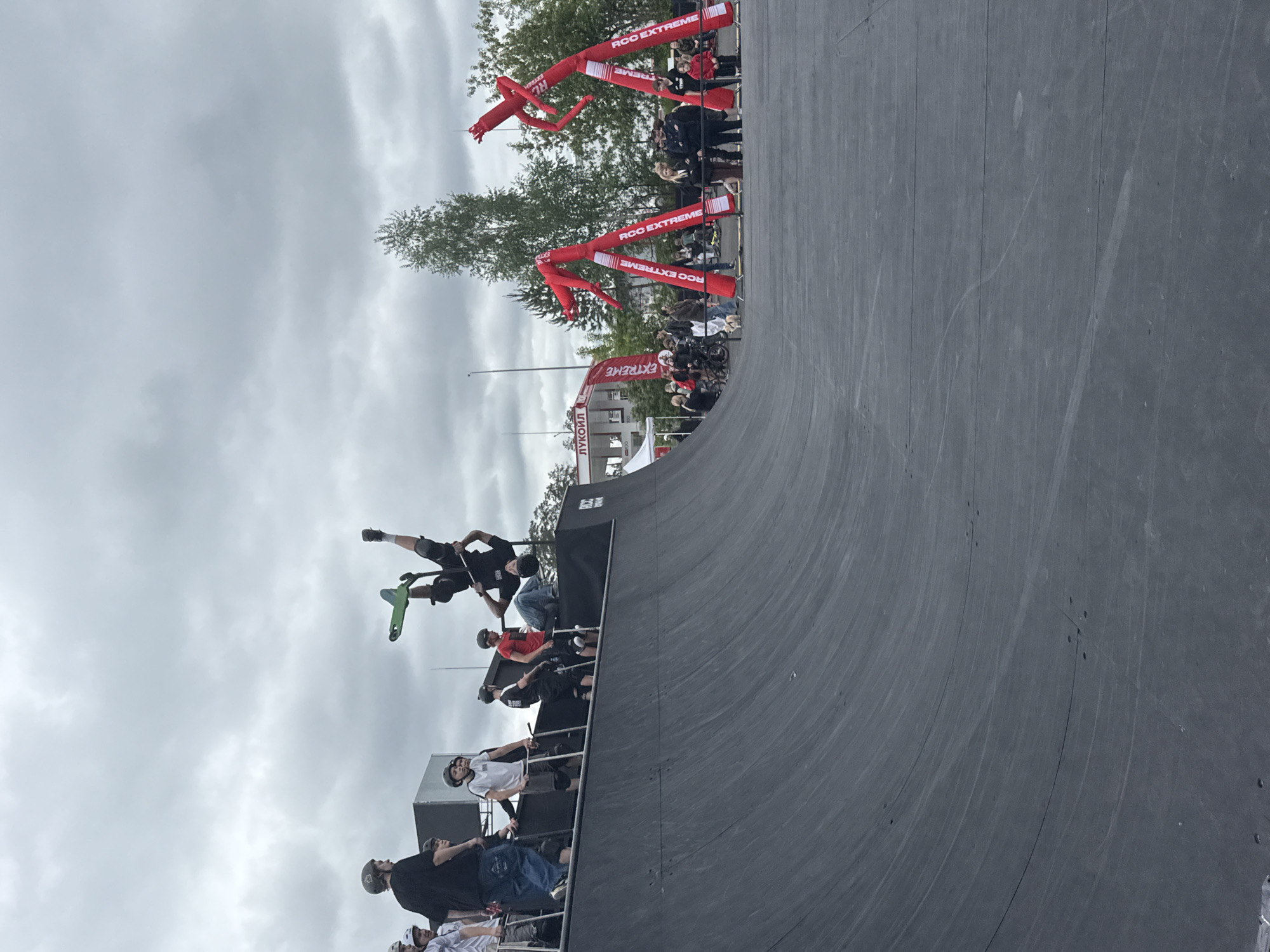 A vertical, wide shot depicts a man mid-air on a green scooter, executing a trick above a vert ramp. A row of spectators line the top edge of the ramp, wearing helmets and observing the action. In the background, red inflatable tube dancers with the words "ROC EXTREME" are visible, along with a crowd of people. A flag also displaying "EXTREME" is present, as well as a building labeled "ЛУКОЙЛ". The cloudy sky overhead suggests an overcast day. Based on the brand names written in Cyrillic, the location is likely in Russia, specifically Yekaterinburg given the provided information.