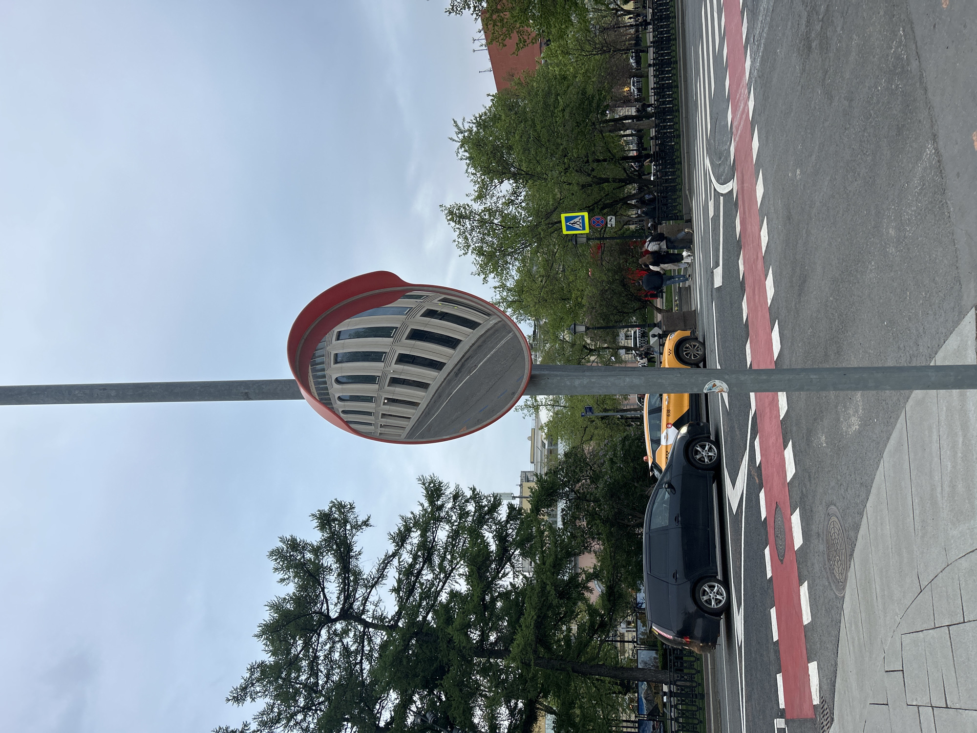 A street scene is captured with a convex mirror in the foreground reflecting a building. The mirror, framed in red, is mounted on a gray pole. Below the mirror is a tall tree with green foliage.
Adjacent to the street is a sidewalk with a black metal fence. A dark-colored car is parked next to a yellow taxi cab on a road marked with red and white stripes. Further down the street, a pedestrian crossing sign is visible with people walking on the sidewalk. The sky is overcast.