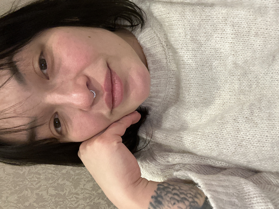 The image presents a close-up, indoor portrait of an adult female in Chaikovsky, Russia. The subject has dark, short hair with bangs and brown eyes, looking directly at the camera with a subtle smile. She wears a silver-colored septum piercing adorned with a row of small, round studs. Her left hand is raised, with her chin resting on her knuckles, and a portion of a dark-ink tattoo is visible on her left forearm, extending from under the sleeve of her light-colored, textured knitted sweater. The background is a blurred, light-toned wall covered in wallpaper featuring a subtle, repeating patterned motif.