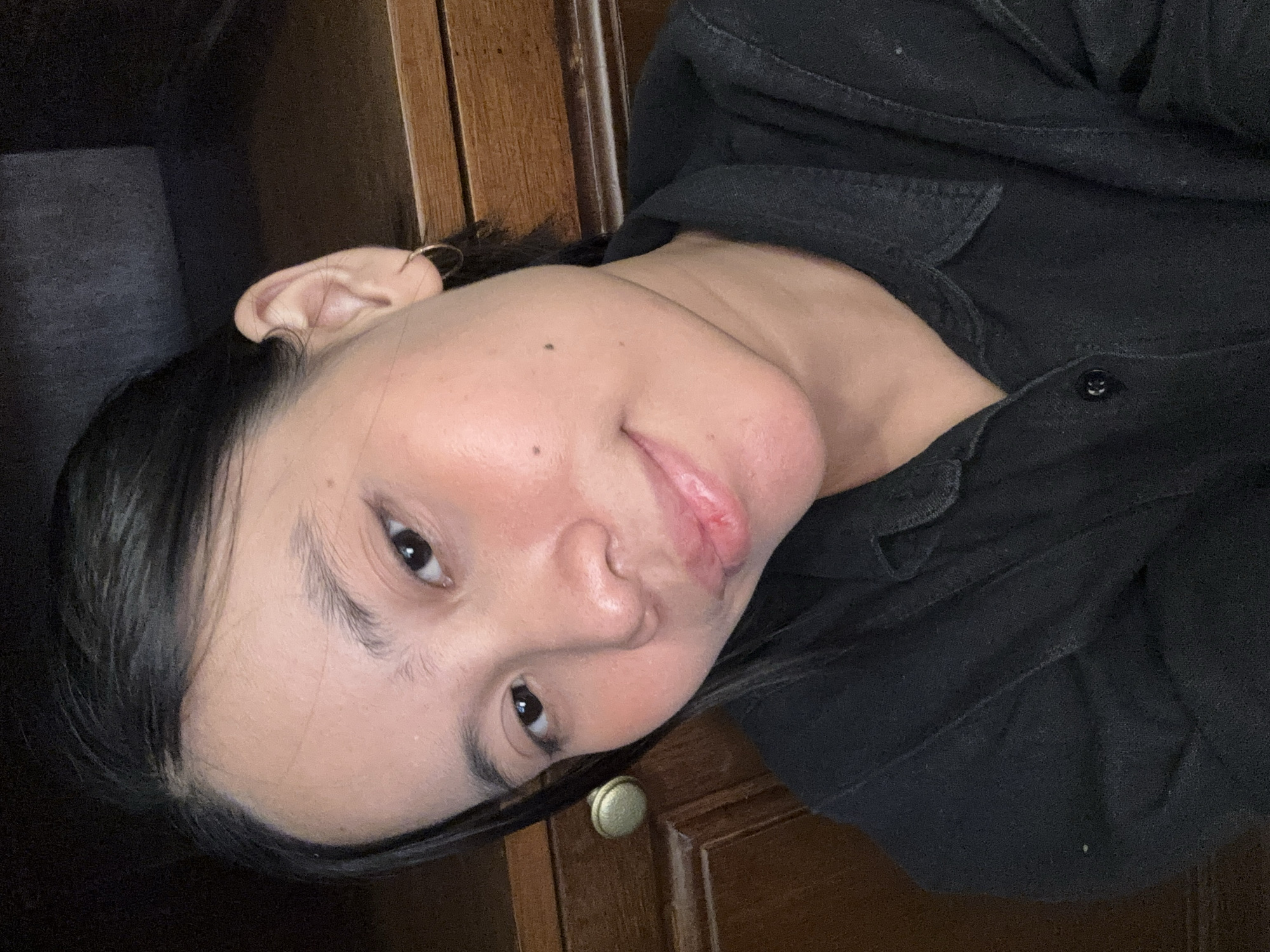 A close-up, head-and-shoulders selfie of an East Asian woman is shown. She is positioned to the right of the frame, with her head tilted and her gaze directed slightly off-camera to her left. Her dark hair is pulled back, revealing her left ear, which has a small hoop earring. Her complexion is light with a reddish blush on her cheeks. She is wearing a black collared shirt. The background consists of a wooden cabinet with a brass knob visible on the left side of the frame. The lighting appears to be artificial and dim, creating shadows. The image is a self-portrait, likely taken indoors in the Kebayoran Baru subdistrict of South Jakarta, Indonesia, given the style of the cabinet and the typical residential interior of the area.