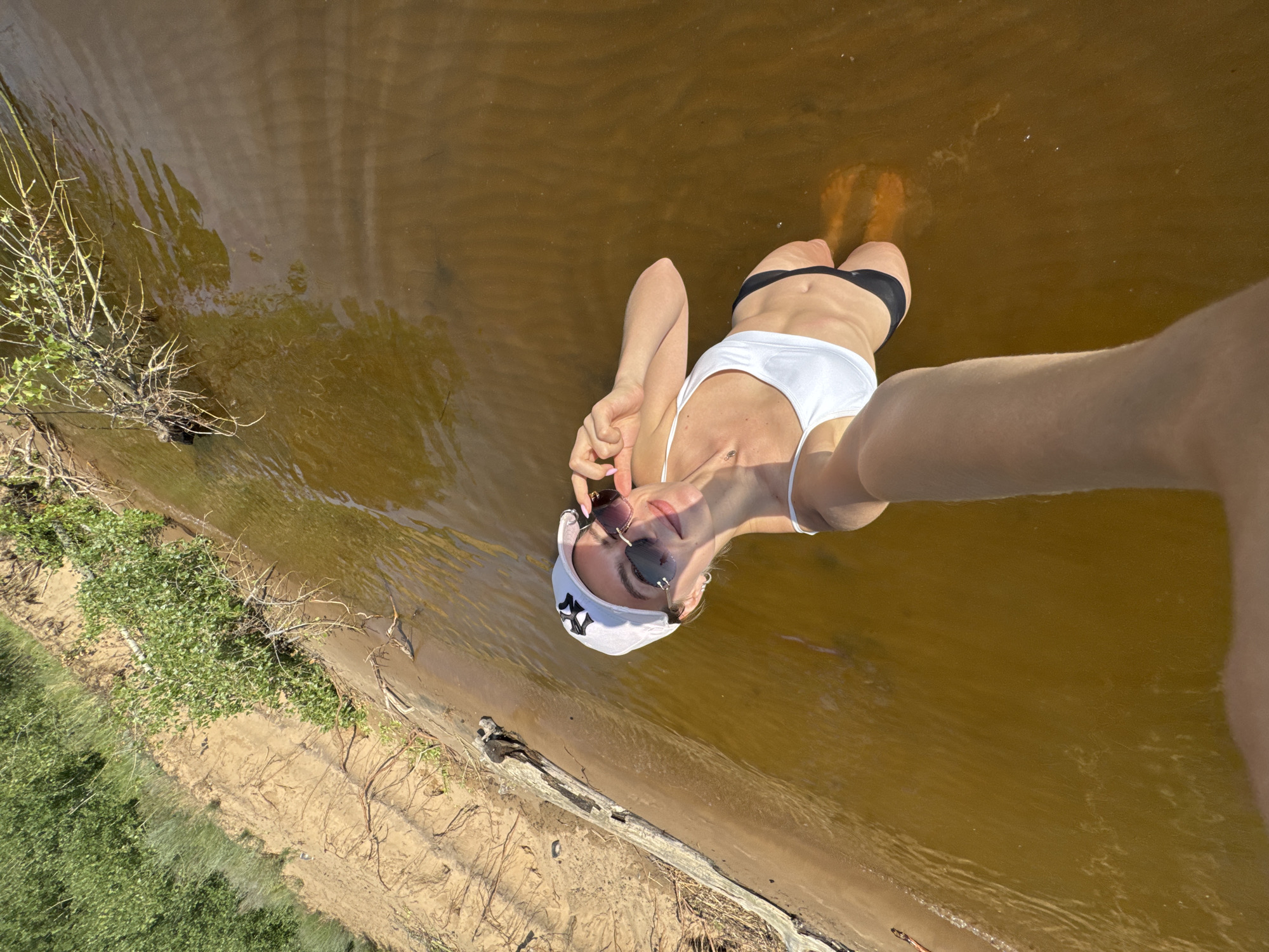A young woman is standing in shallow, muddy brown water, taking a selfie. She is wearing a white bikini top, black bikini bottoms, a white baseball cap with a black logo, and sunglasses. Her right hand is raised, adjusting her sunglasses. The water is calm with small ripples. The background shows a sandy bank with sparse green vegetation and some dead branches. The lighting suggests it is daytime, likely late afternoon due to the long shadows. The location appears to be a natural body of water, possibly a lake or river, in an undeveloped area.