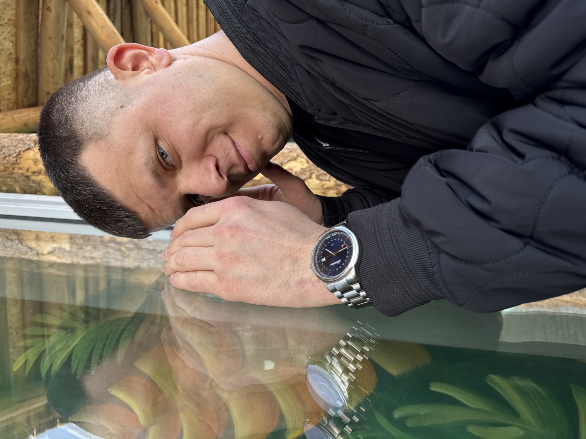 A man is leaning over a glass surface, likely a display case or window, with a wooden structure visible in the background. The man is wearing a dark jacket and a wristwatch with a metal band. The glass surface reflects his image and a leafy design. The location is Perm, Russia.