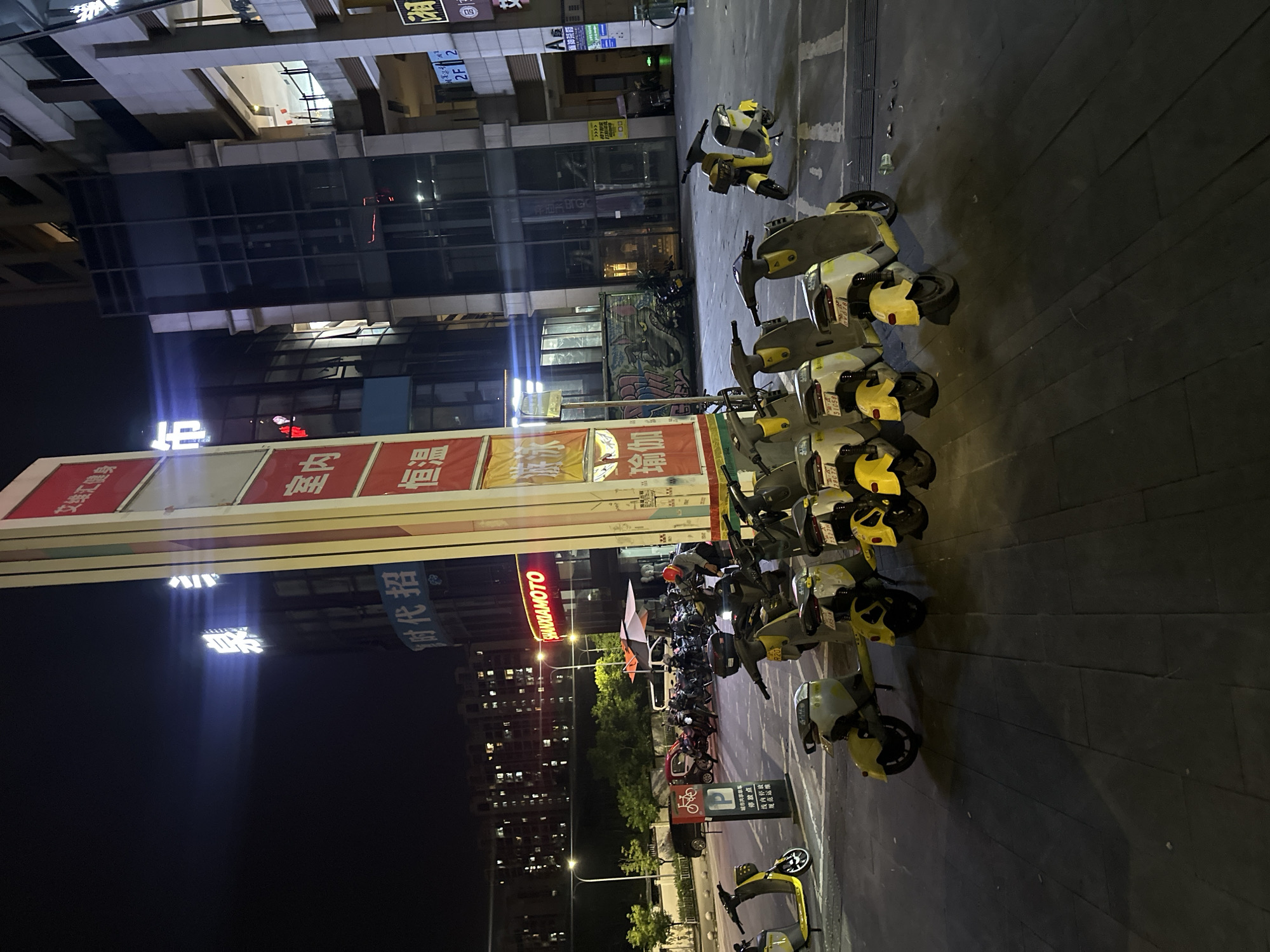 At night, a row of shared electric scooters is parked on a paved area in front of a building. The scooters are predominantly yellow and white. In the background, a tall, illuminated sign displays Chinese characters. The building behind the scooters has a modern glass facade. Further back, apartment buildings with lit windows are visible. The scene suggests an urban environment in the Chongqing High-tech Zone, China.