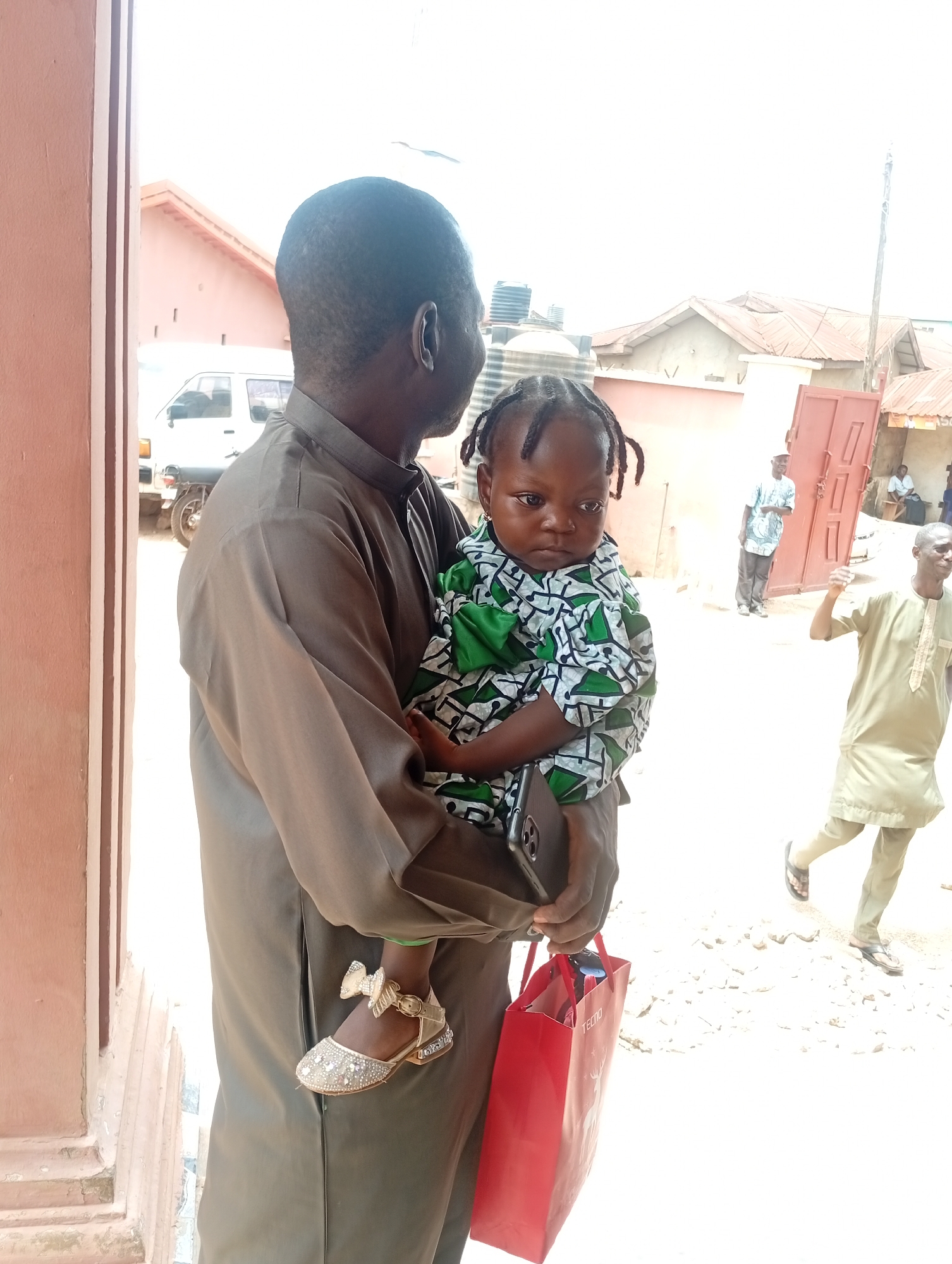 The scene depicts an outdoor street or open area in the city of Sabon Tasha, Nigeria, under bright daylight. In the foreground, a dark-skinned man is positioned with his back and right side towards the viewer, facing slightly to the right. He wears a long-sleeved, collared, olive-green or light brown traditional tunic. He is holding a young, dark-skinned child, who wears a green, white, and black patterned shirt and ornate gold-colored shoes with a bow detail. The child looks towards the left with a neutral expression, hair styled in small braids or twists. The man holds a silver-backed smartphone in his left hand and a red shopping bag with "TECNO" branding in his right hand.
To the left of the man, a portion of a light reddish-brown building with vertical architectural features forms the edge of the frame. In the mid-ground, a white utility van is parked behind the man, with a partial view of a motorcycle wheel behind it. Beyond this, several single-story or low-rise buildings with light-colored walls and reddish-brown corrugated metal roofs are visible, some with black water tanks on top.
On the right side of the frame, a dark-skinned man in a light-colored traditional tunic and pants walks towards the right, away from the camera. Further back, another dark-skinned man wearing a light blue or green patterned shirt stands near an open reddish-brown door. The ground appears unpaved and dusty. The background is overexposed, indicating strong sunlight, and a tall, thin utility pole extends upwards on the far right.