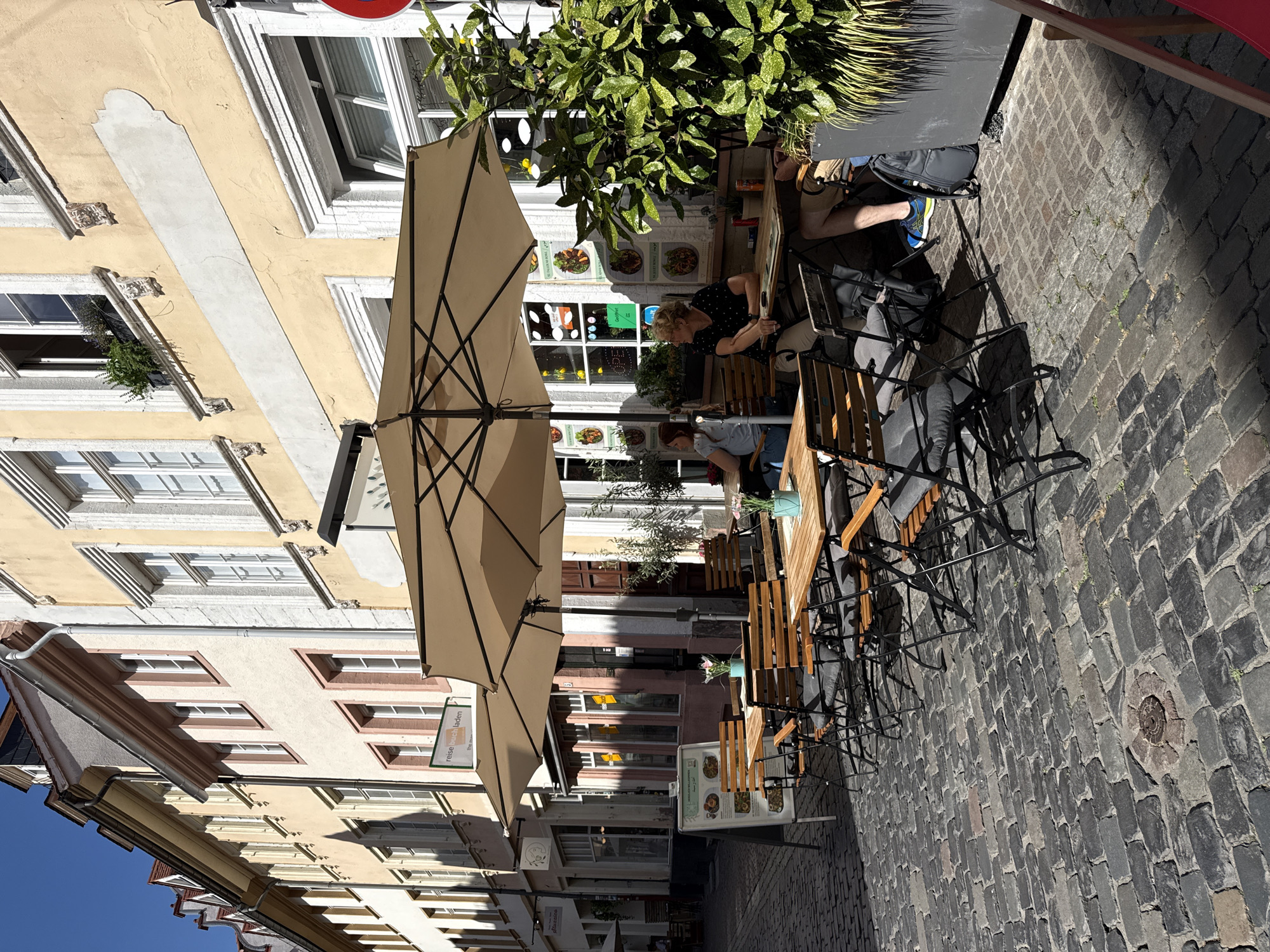 The image depicts an outdoor seating area of a cafe or restaurant on a cobblestone street in Heidelberg, Germany. The cafe is situated in front of buildings with traditional European architecture, characterized by light-colored facades, large windows, and ornate detailing.
Several wooden tables and chairs are arranged under a large, beige umbrella, providing shade. Some tables are set with placemats and a small potted plant. On one table, a menu board is visible. Two women are seated at a table, one looking towards the camera and the other looking away. Another person's legs and feet are visible at a table to the right, wearing blue and yellow athletic shoes and shorts, with a backpack nearby.
A large green potted plant in a dark gray planter is positioned to the right of the seating area, partially obscuring the building behind it. Signs are visible on the building, including one that appears to be a menu with images of food. The street itself is paved with dark, irregular cobblestones, and a circular metal grate is set into the ground in the foreground. The scene is bathed in bright sunlight, casting distinct shadows.