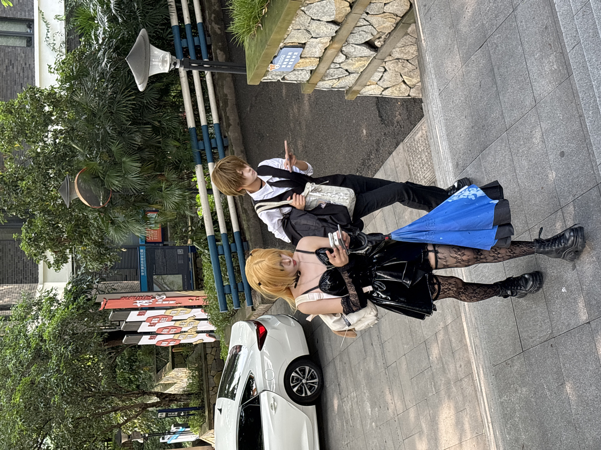 The image captures two individuals in costume on a street in the Yuzhong District of Chongqing, China. The environment includes sidewalks, street lamps, trees, informational signs, and a white car. One person is dressed in a black and white uniform-like outfit, carrying a bag, and gesturing with their hand. The other is in a black patent leather outfit with lace stockings and black boots. They are holding what appears to be a phone. The white car is parked near them.