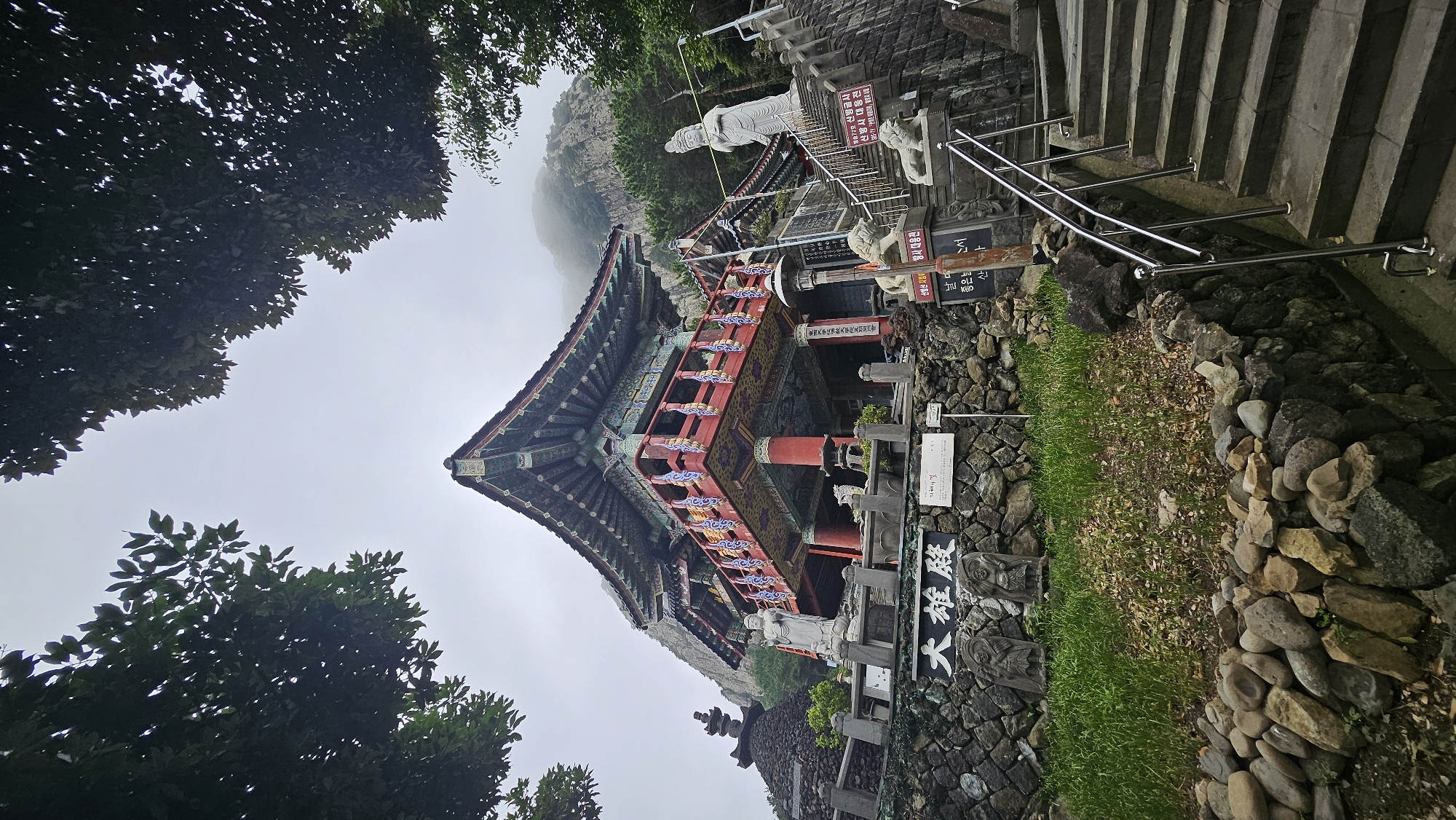 The image depicts a Buddhist temple structure located on a mountainside in Seogwipo-si, South Korea. The temple, painted in vibrant red and blue, features an ornate, multi-tiered roof and a balcony adorned with intricate carvings. In front of the temple, a stone staircase with metal railings ascends. Several stone statues, including a large standing Buddha and two guardian lions, are visible. A stone wall with inscribed Korean characters runs along the lower portion of the scene. Lush green grass and scattered rocks surround the base of the structure. The background reveals a misty, rocky mountain range, indicating the temple's elevated and somewhat remote location. The sky is overcast.