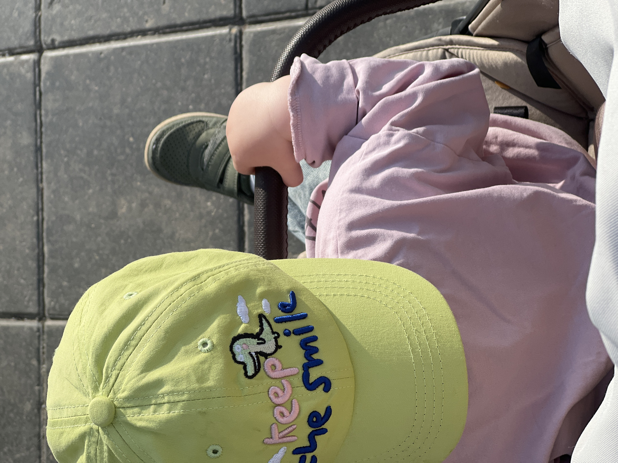 A high-angle shot shows a child in a stroller. The child wears a pink sweater and has a green shoe visible near the stroller's handle. A light green baseball cap with the phrase "Keep the Smile" is in the foreground. The background consists of grey pavement. The location is Khimki, Russia.