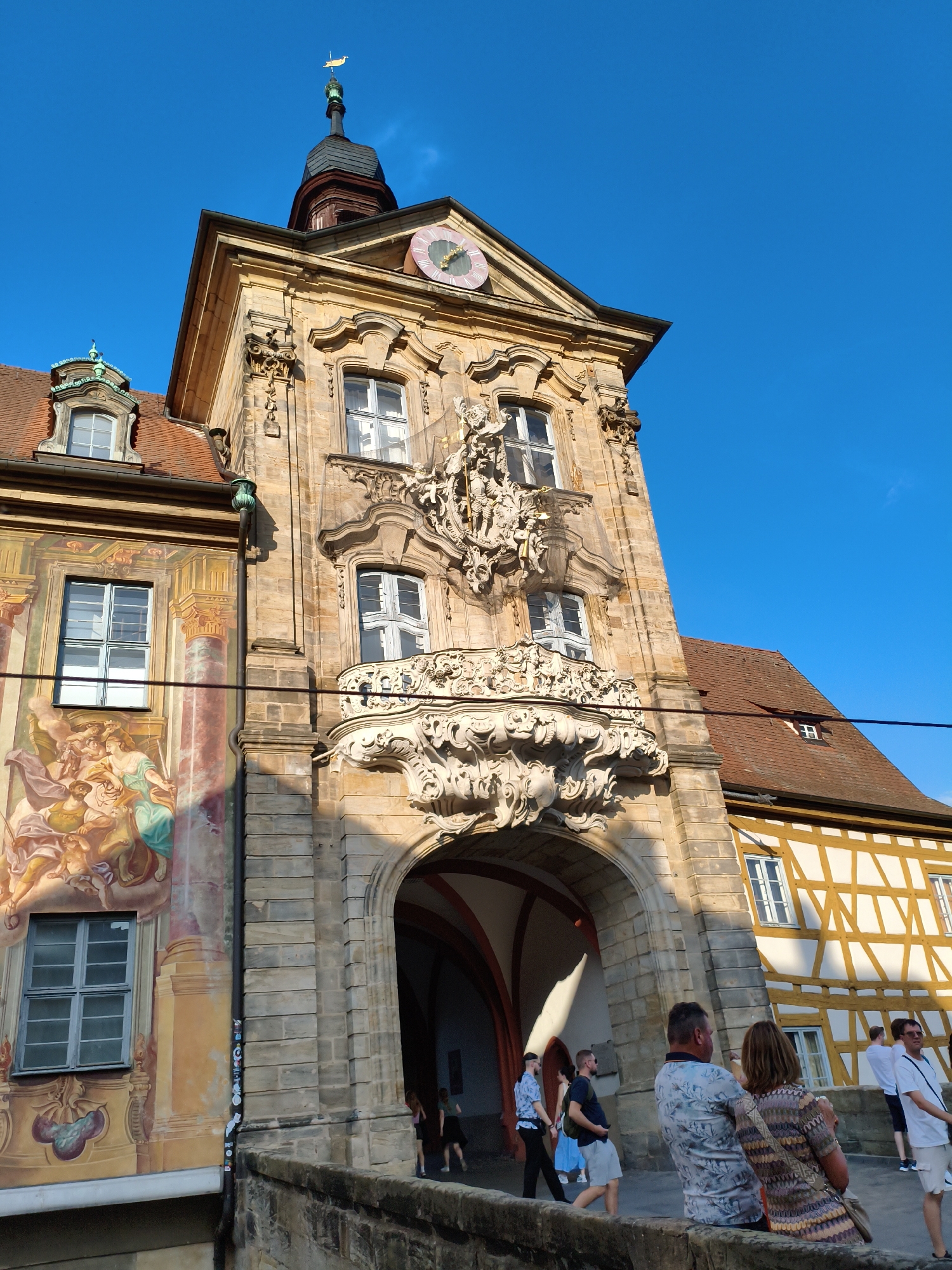 This is a low-angle, medium shot of the Old Town Hall (Altes Rathaus) in Bamberg, Germany, on a sunny day with a clear blue sky. The building, constructed from a warm, tan stone, features intricate Baroque architectural details. A prominent archway leads into the building's interior, with a balcony adorned with elaborate white stucco carvings situated above it. A large clock with a pink face is visible on the facade. To the left of the main building, a section of the wall is decorated with a colorful fresco depicting several figures in flowing drapery. Windows are visible on various levels of the building, some with white panes and others with a light blue hue. A red-tiled roof crowns parts of the structure. To the right, a building with a distinctive half-timbered facade, featuring white infill between dark wooden beams, is partially visible. In the foreground, a low stone wall separates the viewer from the building. Several people are present, some walking along the wall and others gathered near the archway. A couple is seated on the wall, with the man's arm around the woman's shoulders. Another man stands to their right, facing away from the camera. A group of people is visible walking through the archway. The scene is bathed in sunlight, casting shadows.