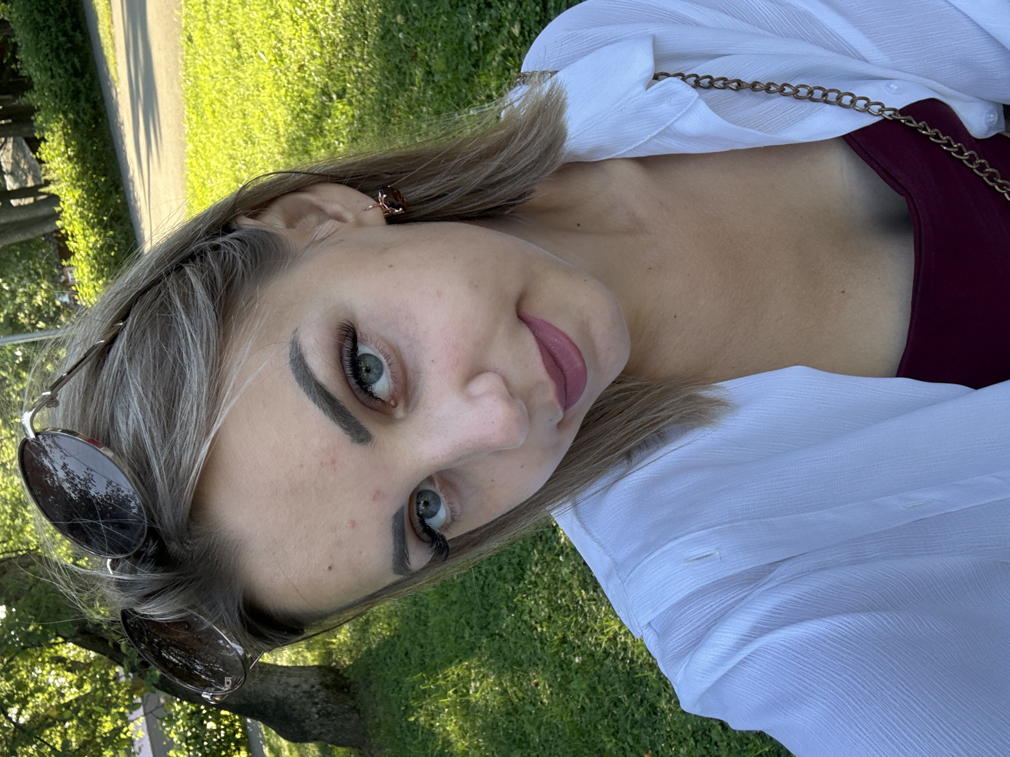 An outdoor, eye-level, medium shot captures a young woman taking a selfie in a park during daylight. She is positioned in the center of the frame, looking directly at the camera with a slight head tilt. Her blonde hair is styled down and partially obscured by sunglasses perched on top of her head. She is wearing a white, open-collared, long-sleeved shirt over a dark red top and a gold-colored chain strap is visible. Her makeup includes eyeliner, mascara, and dark lipstick. A single, small, dark earring is worn on her right ear. The background consists of lush green grass, trees, and a paved pathway, suggesting a park setting. The overall lighting indicates it is a sunny day, casting shadows on the grass and highlighting the subject. The location is identified as Yuzhno-Sakhalinsk, Russia.