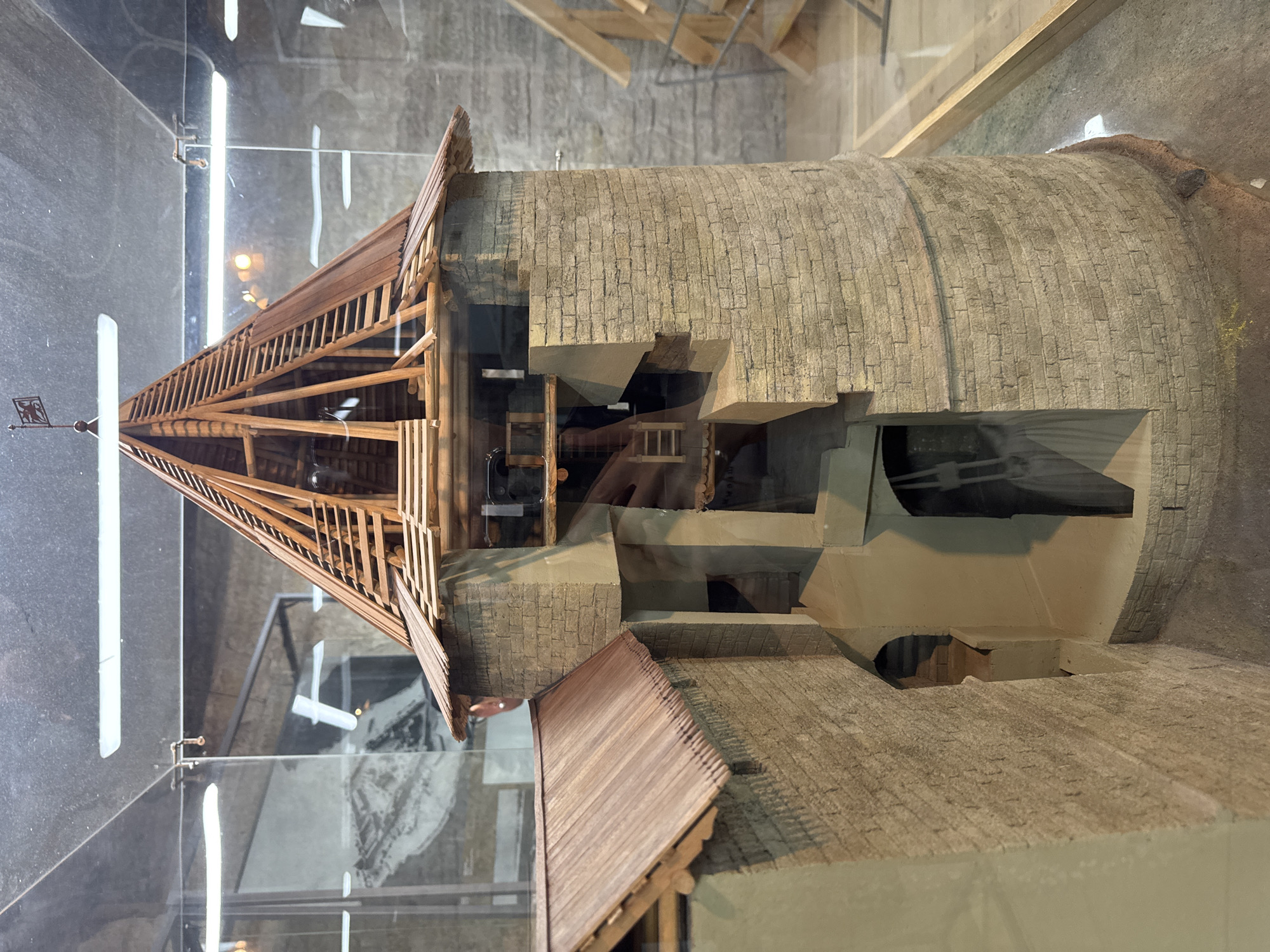 This media file captures a detailed architectural model, likely of a historical structure in Shlisselburg, Russia. The primary subject is a model of a circular tower or building with a conical wooden roof, constructed from what appears to be brickwork. The model is displayed within a glass enclosure, indicated by reflections on the surface.
The tower has several openings and recessed areas, suggesting internal structures or stages within the building. Part of the roof is missing or incomplete, revealing the wooden supports and beams beneath. A prominent wooden staircase or ramp spirals up the exterior of the tower, leading towards the conical roof. At the apex of the roof, there's a small decorative element resembling a weather vane.
The surrounding setting of the model is a dimly lit indoor space with a concrete or stone-like backdrop. The lighting appears to be artificial, casting shadows and highlighting the textures of the model. The overall presentation suggests it is part of a museum exhibit or a historical diorama. No people are visible in the frame, and the scene is static, representing a fixed architectural subject. The time of day and weather are irrelevant as it is an indoor display. There is no visible text in the image.