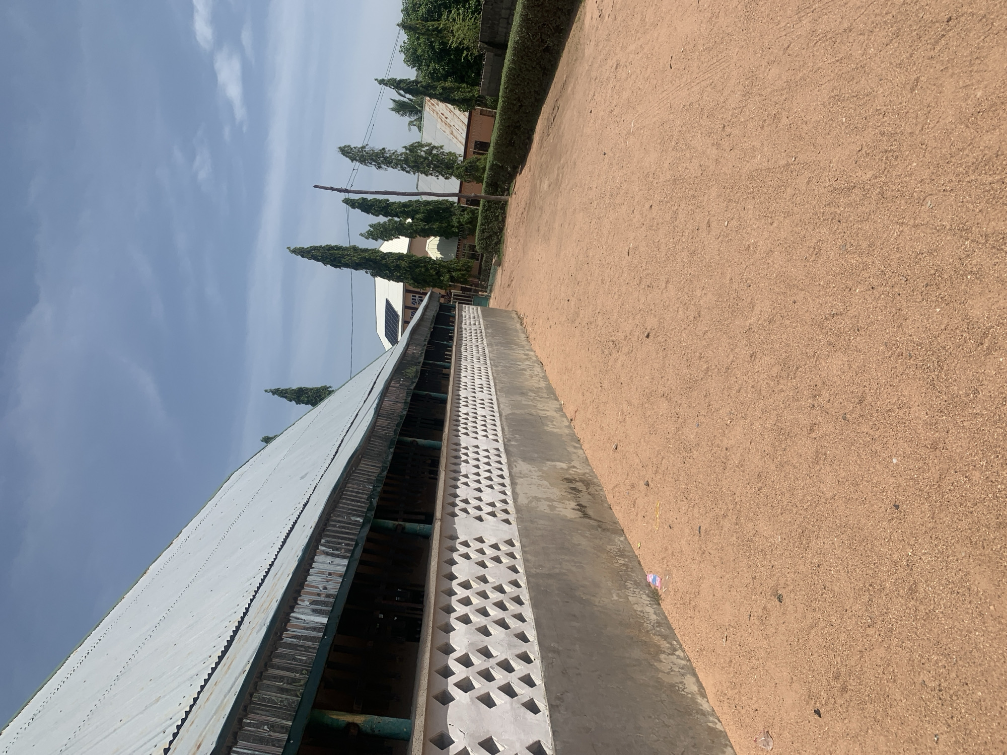 The image depicts an outdoor scene in Jauro Yinu, Nigeria, characterized by a dusty, light brown dirt path leading away from a building with a corrugated metal roof. The building, visible on the left side of the frame, features a textured concrete wall with a patterned opening and a dark green overhang. Tall, slender trees are interspersed throughout the background, some partially obscured by other structures. Several other buildings with similar roofing materials are visible in the distance. The sky is a pale blue with scattered white clouds. A few pieces of plastic litter are present on the ground near the building. There are no people visible in the image.