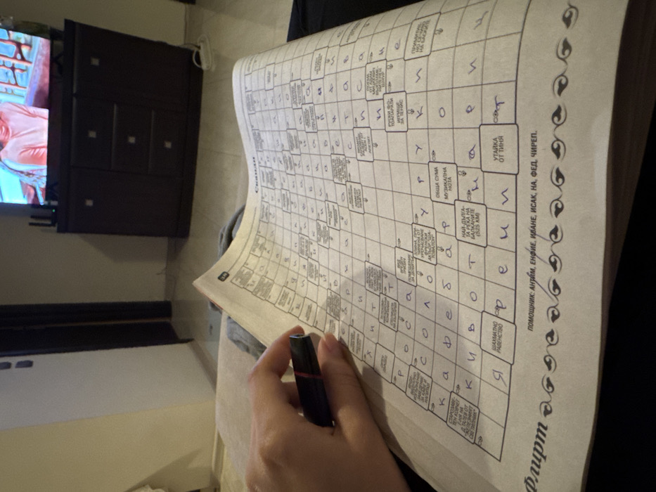 A person's left hand, with light skin tone, holds a black pen with a red accent over an open, white-paged crossword puzzle book or magazine. The puzzle, titled "Скандал" (Scandal) and featuring "Флирт" (Flirt) at the bottom, is written in Bulgarian Cyrillic, with numerous cells already filled in blue ink. The individual appears to be seated on a light-colored upholstered surface, possibly a sofa. In the background, a dark brown wooden cabinet with multiple drawers supports a flat-screen television displaying a partial image of a person wearing an orange or peach-colored top. The room features light-colored walls, light flooring, and a dark doorway is visible on the left. The Bulgarian language content on the crossword puzzle strongly indicates the scene is located in Bulgaria, most likely the capital city of Sofia.