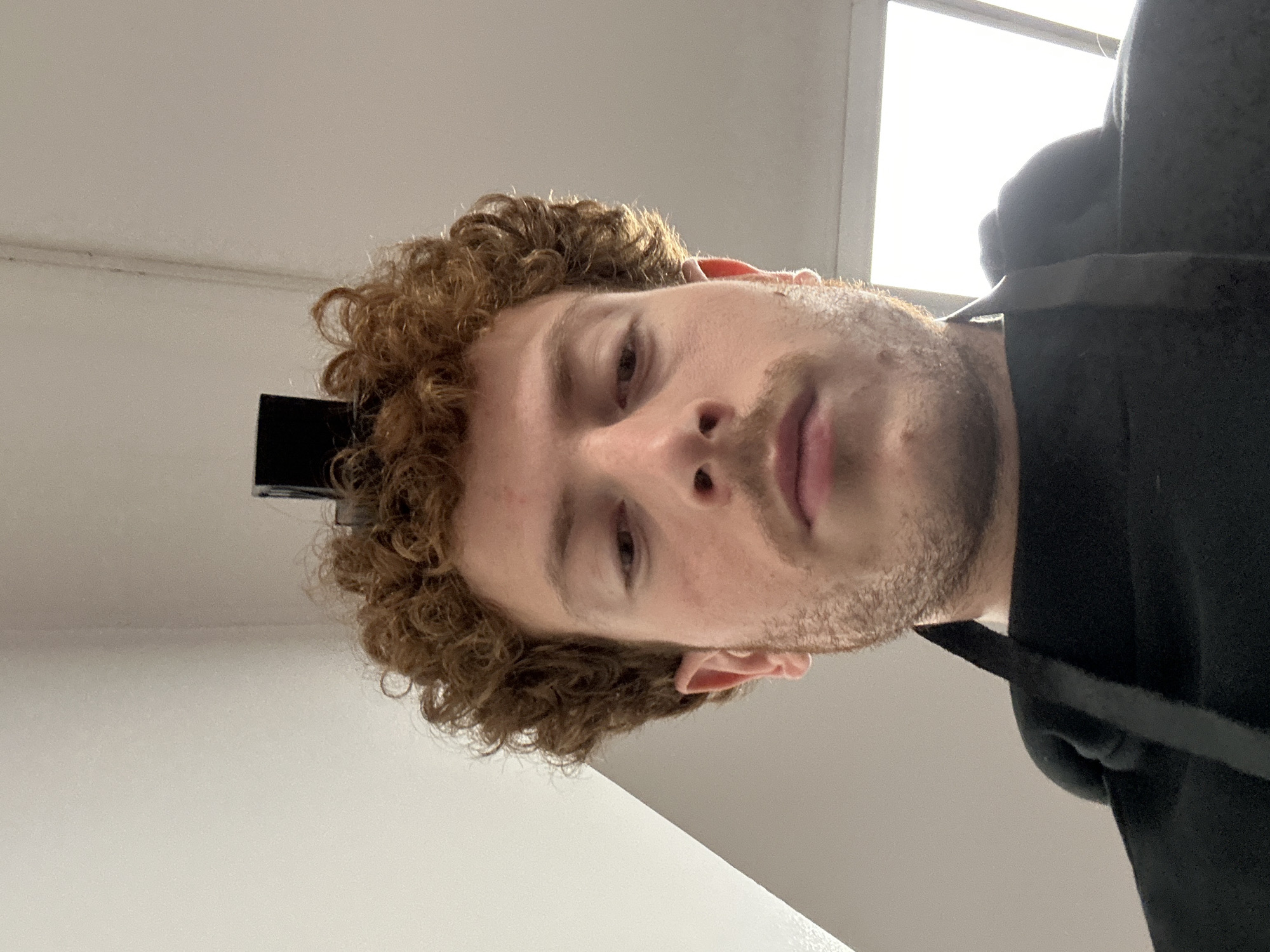 The image displays a close-up, low-angle portrait of an adult male subject in an indoor setting within the city of New York, United States of America.
The subject has light skin, curly reddish-brown hair, a mustache, and short stubble or a very short beard. He is wearing a dark, presumably black, hooded garment, with a black strap visible over his left shoulder, possibly indicating an apron or shoulder bag. A small, dark, rectangular object is positioned upright within his hair on the top of his head. The subject is looking directly downwards, consistent with a selfie perspective.
The background consists of light-colored, plain walls and a ceiling. To the lower right of the frame, a bright, diffuse light source, consistent with a window or skylight, is visible, indicating natural illumination entering the space. No other specific environmental identifiers are present.
