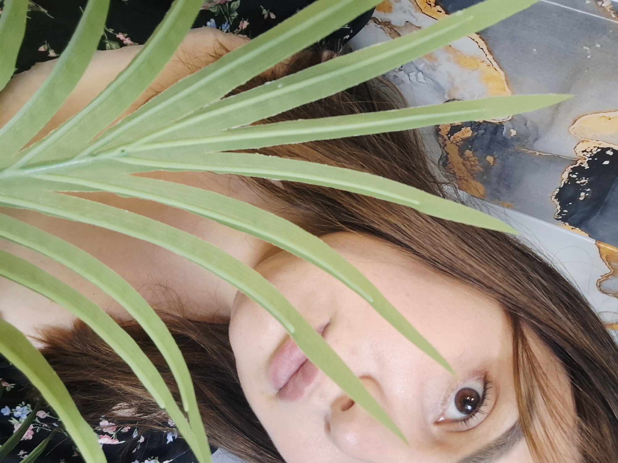 The scene is located in the city of Chaikovsky, Russia. A person, appearing to be female, is positioned behind a cluster of green, elongated plant leaves, which partially obscure her face. Her right eye, featuring a brown iris, is clearly visible, along with her nose and lips, which are closed. Her light-toned skin is evident. She has brown hair, visible along her forehead and on the right side of her head. She is wearing a dark-colored garment adorned with small pink, blue, and white floral patterns, visible on her left side. The background features an abstract, mottled surface resembling marble or stone, characterized by shades of grey, black, and distinct patches of gold or yellow.