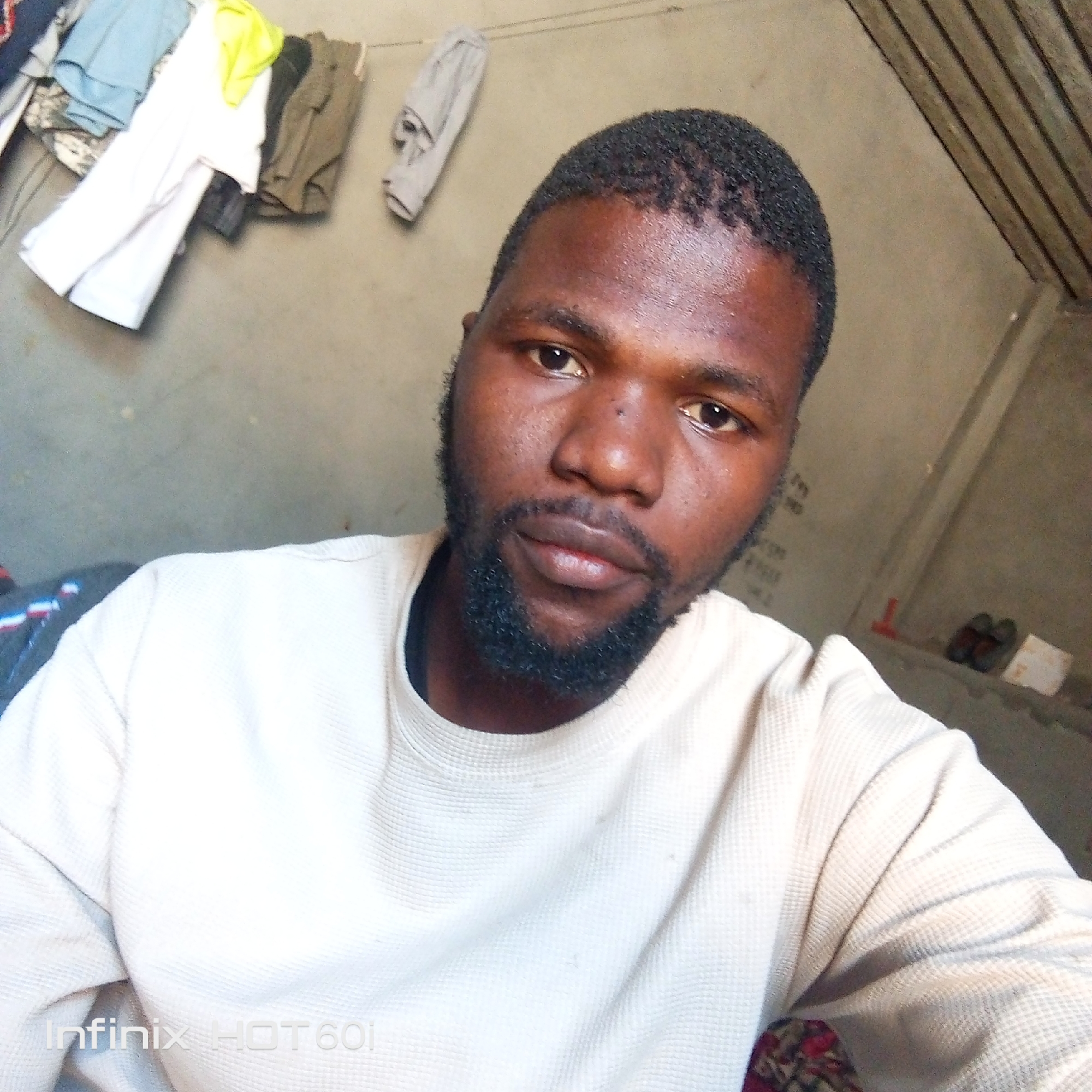 A close-up, eye-level shot of a Black man with short, dark hair and a beard, wearing a light-colored waffle-knit shirt. The background features a pale, undecorated wall with clothes hanging on a line. The ceiling is sloped and has visible beams. In the lower right corner, a pair of shoes and a white object rest on a surface. The bottom left corner shows the watermark "Infinix HOT 601". The location is the city of Unknown, Unknown.