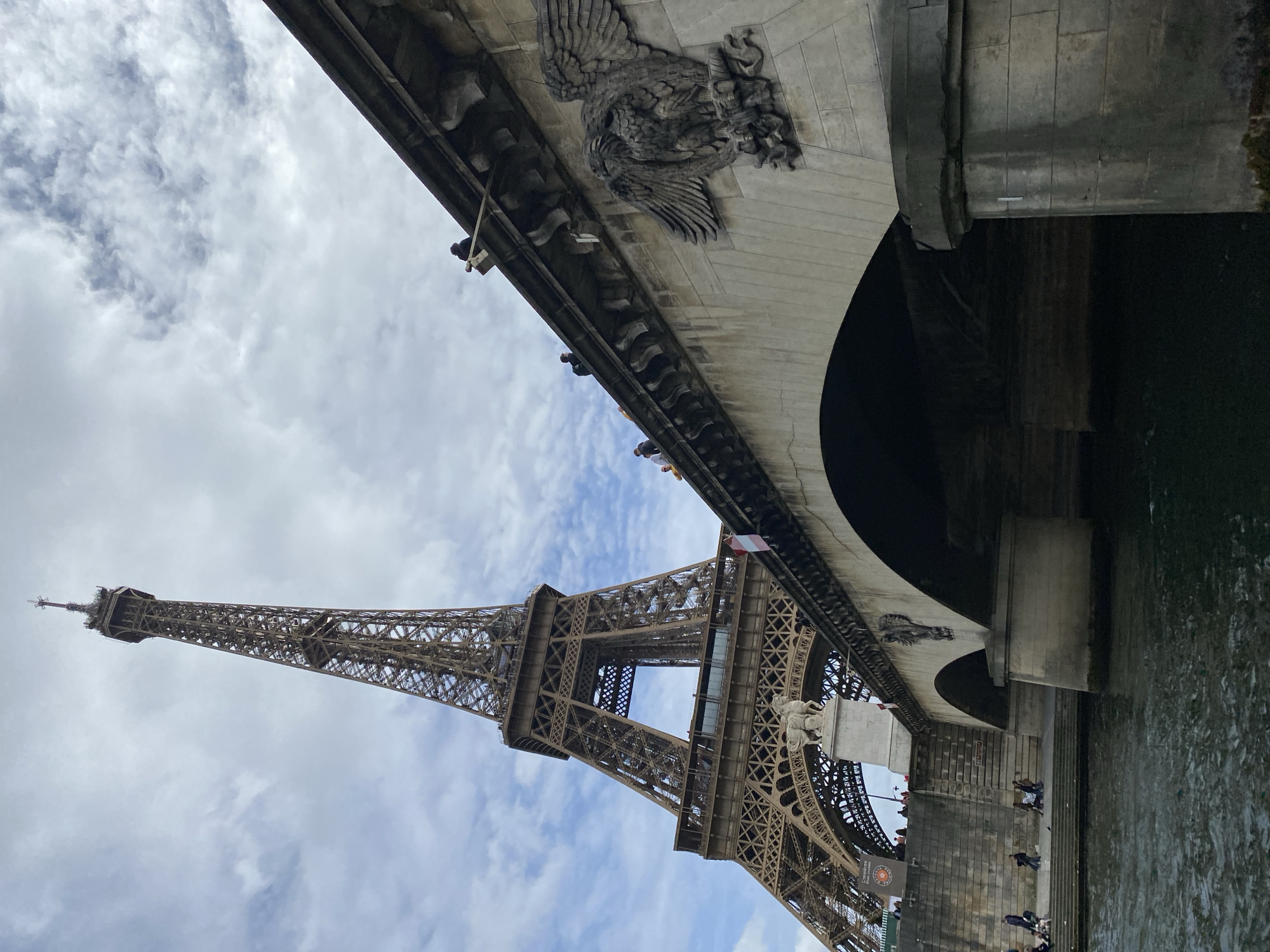 The image shows the Eiffel Tower in Paris, France, viewed from below a bridge spanning the Seine River. The tower rises against a cloudy sky. The bridge features stone architecture, including carved ornamentation. People are visible along the riverbank. The water of the Seine is visible.