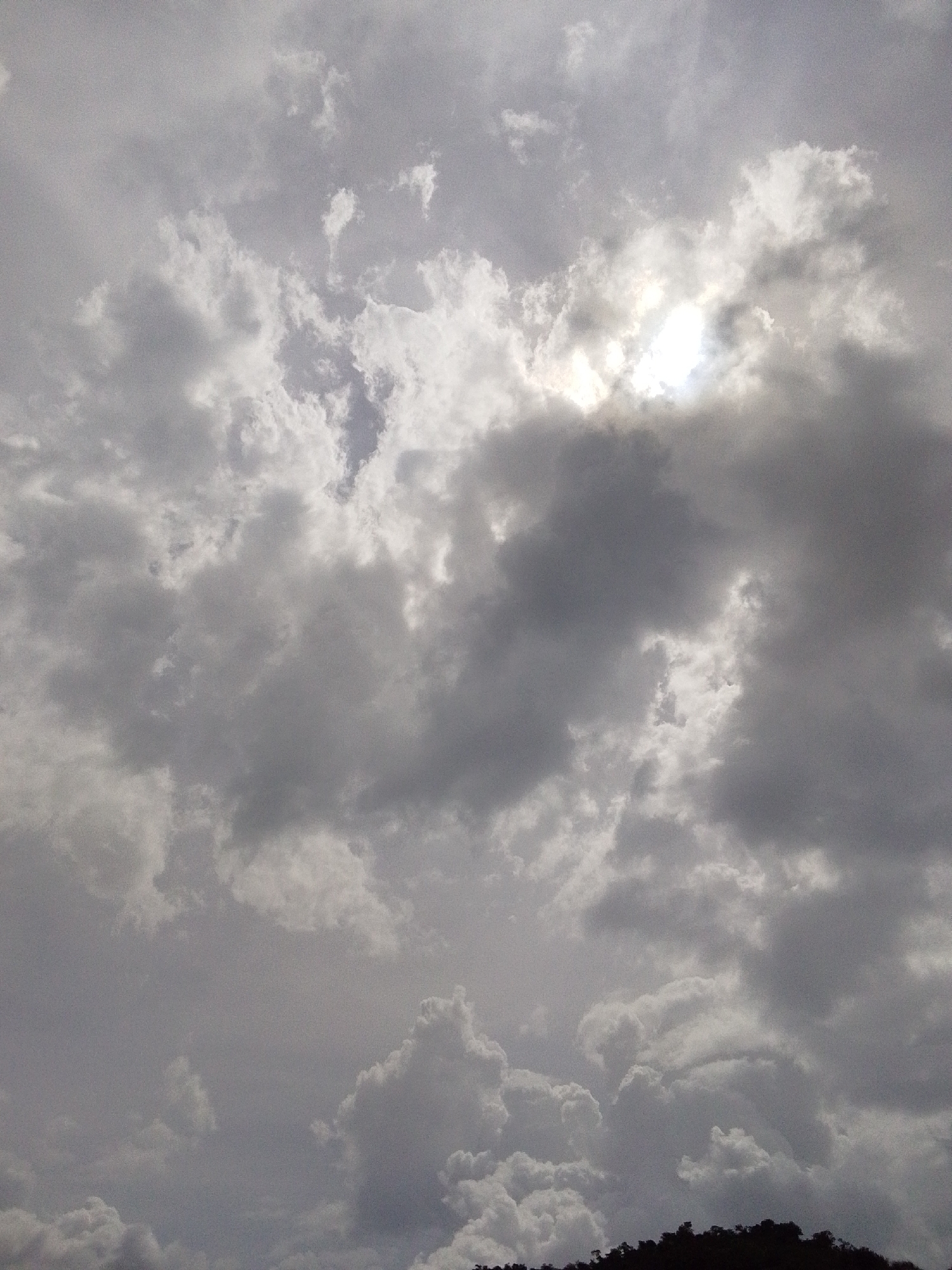 This is a photograph of the sky above Serti, Nigeria, featuring a dramatic cloudscape. The image is dominated by dense, grey clouds, with patches of brighter white clouds interspersed. Sunlight is visible breaking through a gap in the clouds towards the upper right portion of the frame, creating a bright, luminous effect. The bottom of the image shows a dark silhouette of trees, indicating a landscape beneath the sky. No people or specific objects are discernible within the clouds or the silhouetted vegetation. The overall impression is of a cloudy or potentially stormy sky.