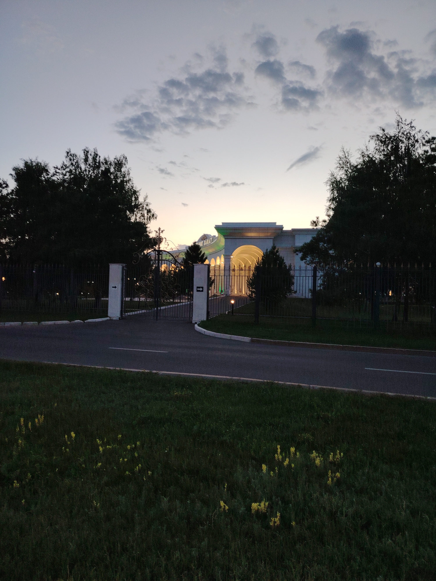 The image depicts the main entrance to the Palace of Peace and Reconciliation in Astana, Kazakhstan, at dusk. The sky is a pale blue with scattered grey clouds, transitioning into a warmer gradient near the horizon. The Palace, a large, light-colored, pyramid-shaped structure, is partially visible in the background, illuminated by internal lights. A grand, arched entranceway with ornate details is the focal point of the building.
In front of the Palace, a black wrought-iron gate stands open, allowing access. The gate is flanked by two white, square pillars, each bearing a black arrow pointing inward. A dark asphalt road leads to the gate, with a white line marking its edge. To the left of the road, a dark metal fence runs along the perimeter, partially obscured by the shadows of tall, dark green trees. Similar fences and trees are also visible to the right of the gate.
In the foreground, a grassy area is sparsely dotted with small, yellow wildflowers. The overall lighting suggests it is either early morning or late evening, with the artificial lights of the Palace beginning to dominate the natural light. There are no people visible in the scene.