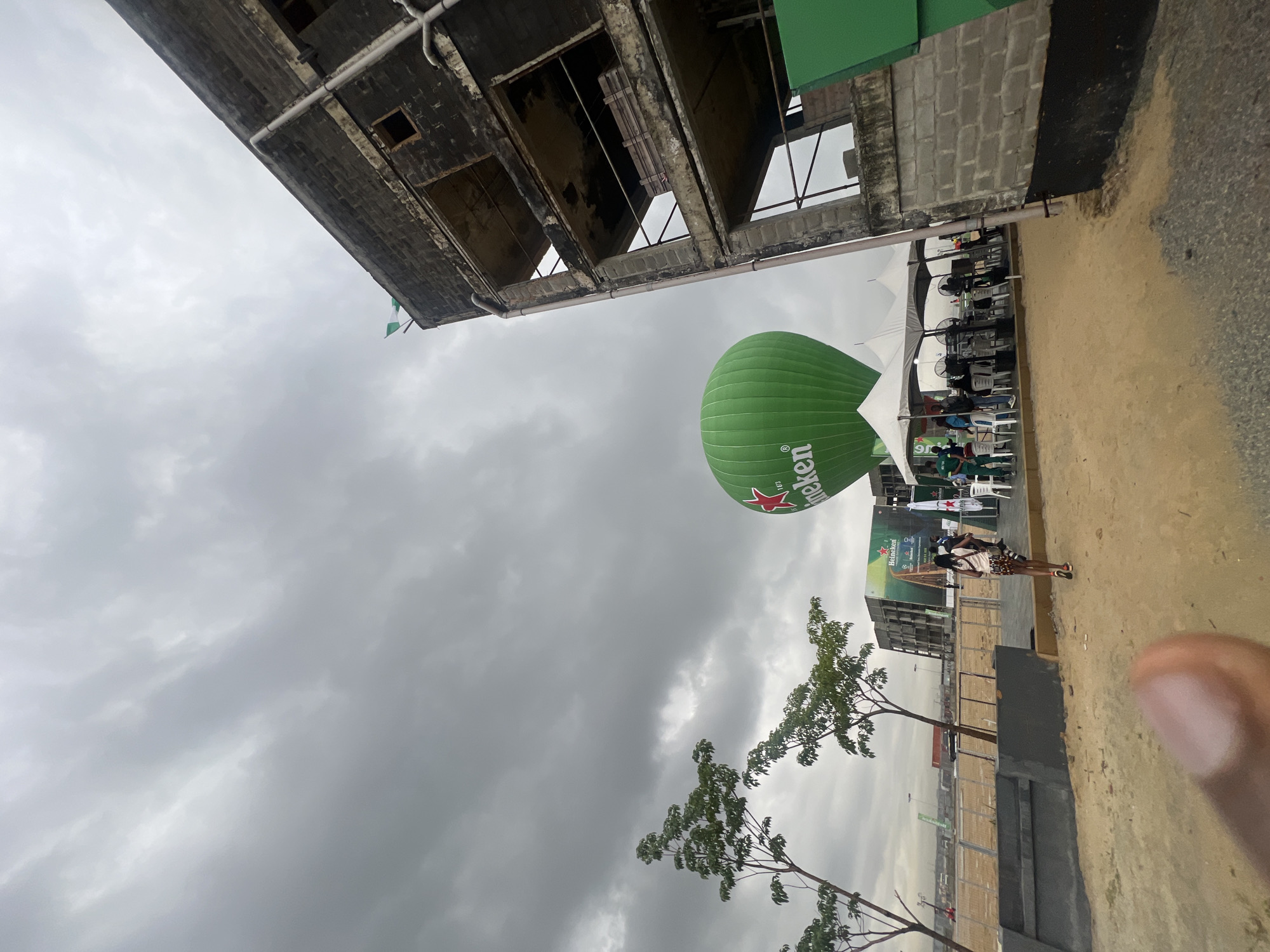 The image, taken outdoors in daylight under a cloudy sky, depicts a scene near Tafawa Balewa Square in Lagos, Nigeria. An incomplete, multi-story building is visible in the upper left. A large, green Heineken hot air balloon floats near tents and seating areas filled with people. A woman stands on a paved area, adjacent to fencing and young trees. A large Heineken advertisement is in the background.