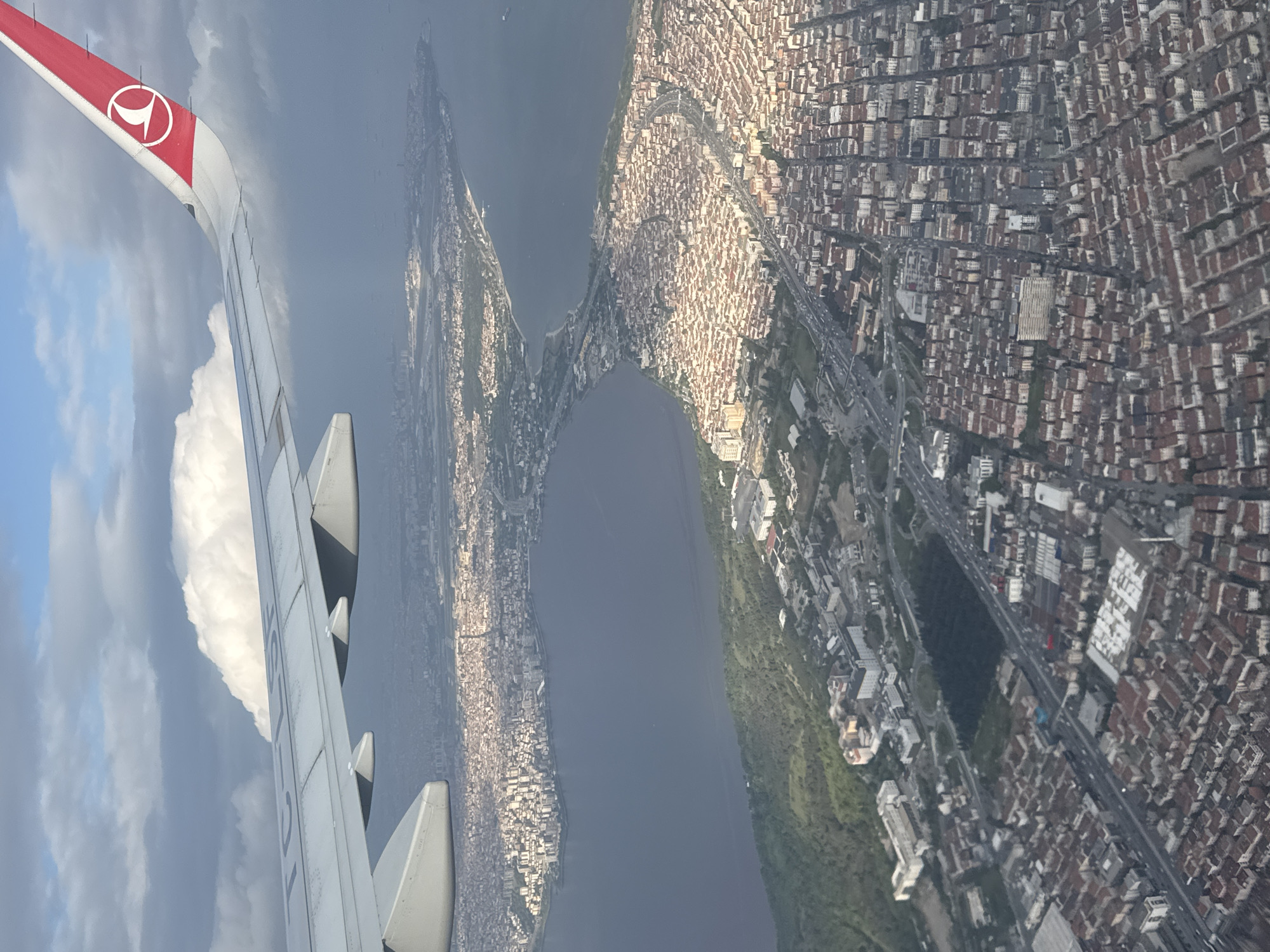 The image is an aerial view from an airplane window, showing a dense urban landscape bisected by a waterway. The wing of a Turkish Airlines plane is visible in the foreground. The landscape on either side of the waterway is heavily populated with buildings, appearing densely packed. Roads run alongside the waterway and through the urban area. The photograph does not appear to show Saint Petersburg.