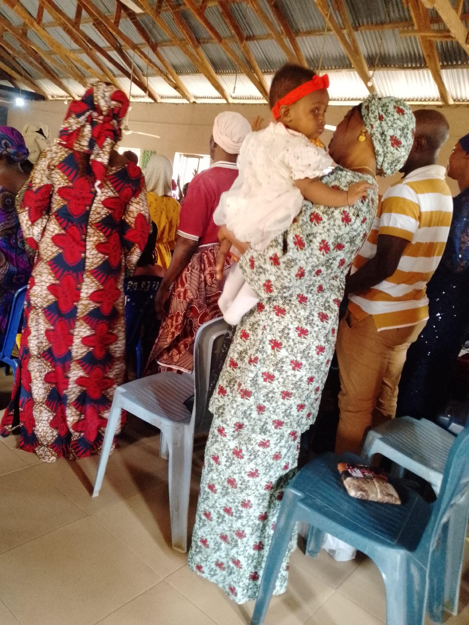 The image shows an indoor scene featuring a group of people, likely in a religious or community setting, given the attire and environment. A woman stands prominently, holding a baby wearing a white dress and a red headband. The woman is dressed in a long, patterned dress with a matching head covering. Other individuals are visible in the background, some wearing traditional African clothing. The environment includes plastic chairs and a building structure with a corrugated metal roof. The location is specified as Unknown, Unknown.