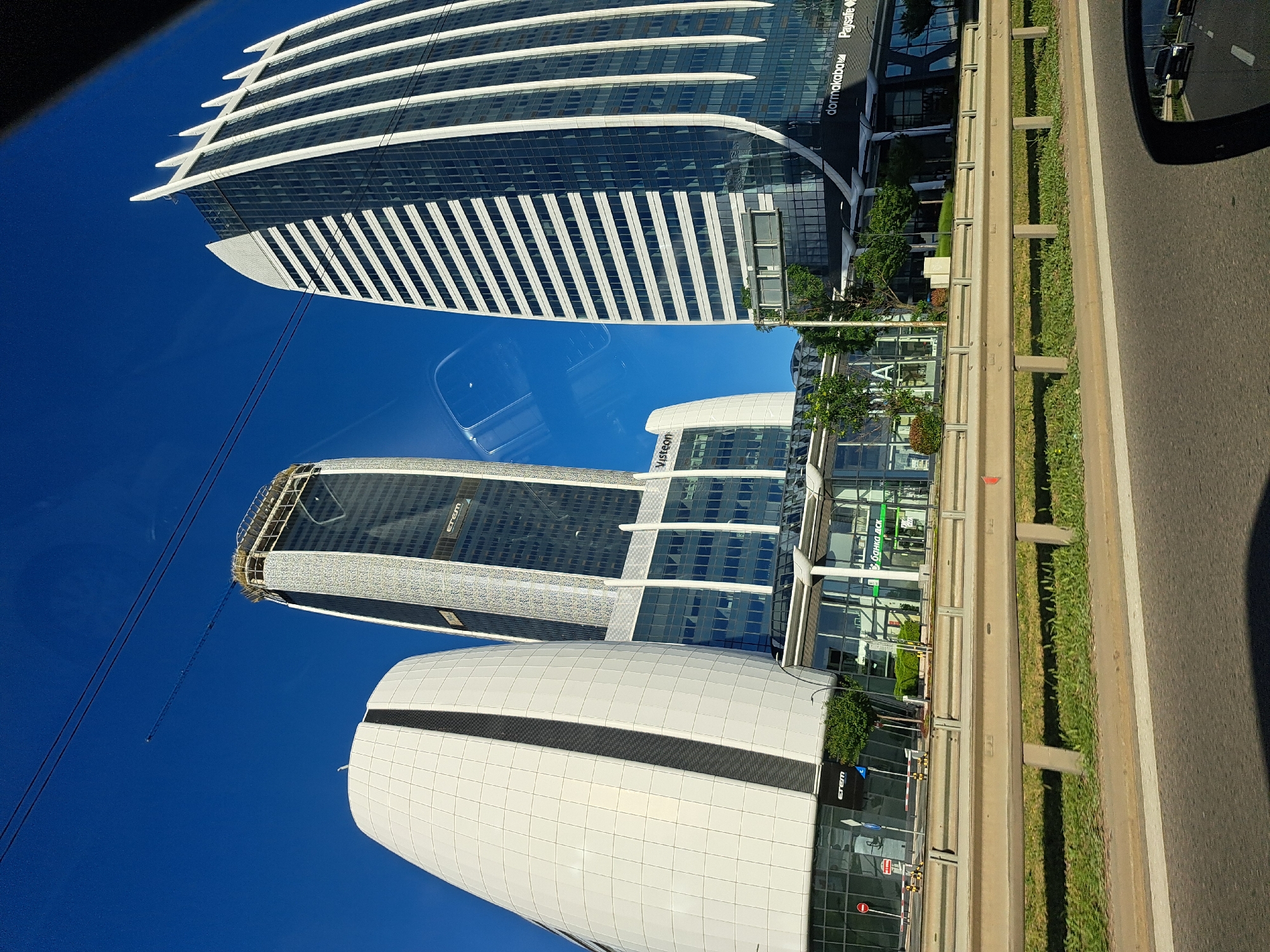 The image shows three modern skyscrapers in the city of Unknown, Unknown. The first skyscraper, at the top, features a white, striped, curved design, and the name "dormakaba Paysafe" is visible on its side. The second skyscraper, in the middle, is a tall, glass structure with a grid pattern and the name "Verison" visible. The third skyscraper at the bottom is white with a dark stripe. Blue sky is visible in the background, and there is a highway guardrail and road to the right of the buildings. The image appears to have been taken from inside a vehicle, as part of the car's interior is visible.