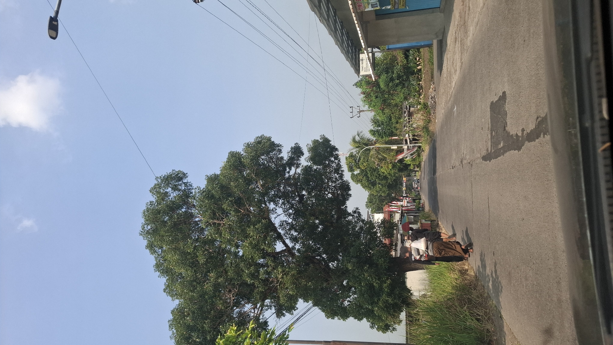 The image depicts a view from a vehicle traveling on a paved road in Sukasari, Indonesia. The road is lined with vegetation on the left and buildings and trees on the right. A large, leafy tree dominates the left side of the frame, with its branches extending over the road. Three individuals are walking on the left side of the road, facing away from the camera. The person in the middle is wearing a light brown sarong and a white shirt, carrying a striped bag over their shoulder. To their left is another person in a black shirt and pants, and to their right is a person in a black hoodie and shorts.
On the right side of the road, a building with a blue door and a gray, sloped roof is visible. A sign on the building reads "TOKO BERAS," indicating a rice shop. Further down the road, there are more trees, buildings, and utility poles with overhead wires. The sky is clear and blue with a few wispy clouds. The road surface shows signs of wear, with patches and some wetness in the center. The overall scene suggests a daytime setting in a somewhat rural or suburban area of Sukasari.