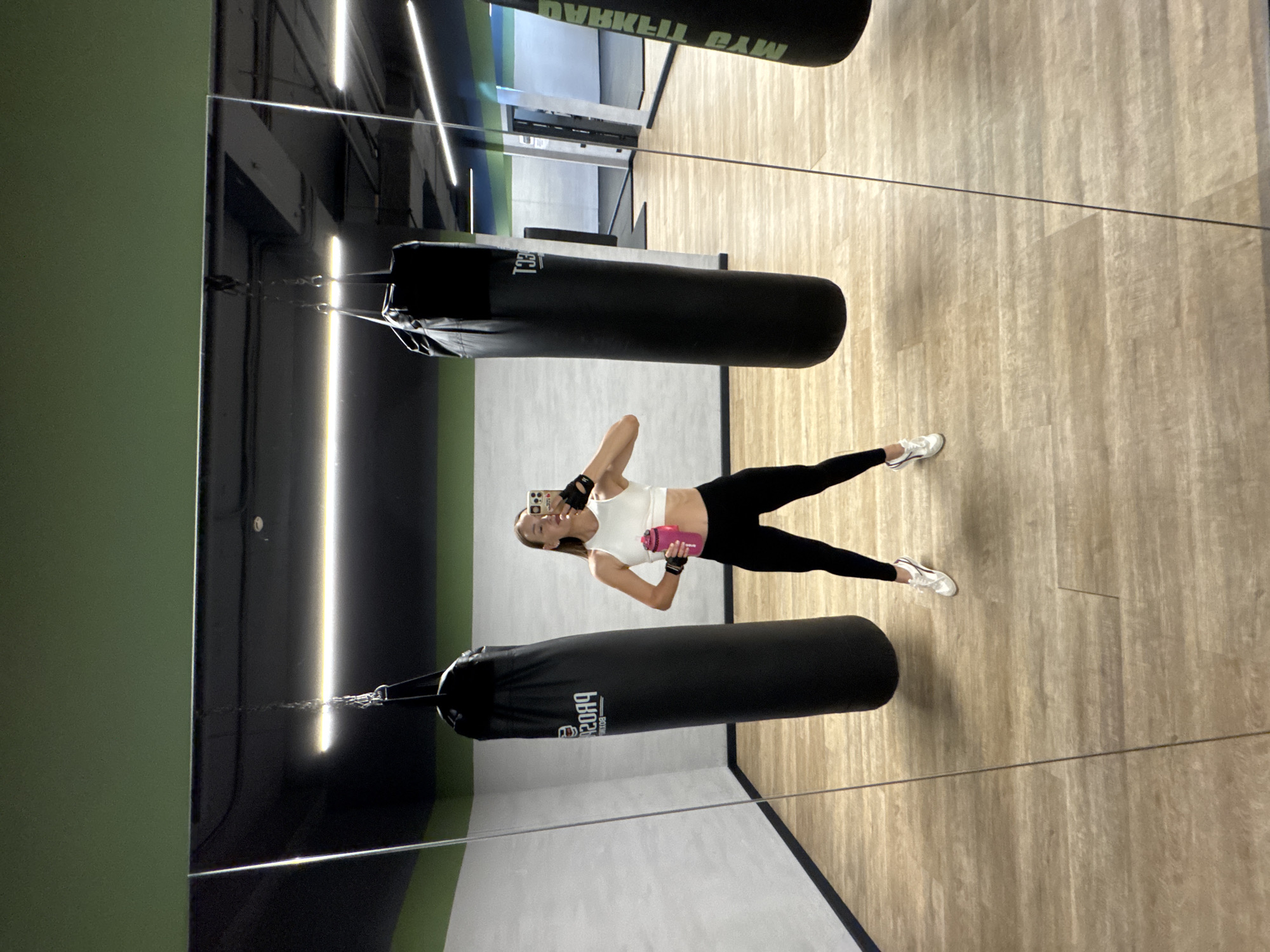 In a gym with a wooden floor and gray walls, a woman in black leggings, a white sports bra, and white sneakers takes a selfie in a large mirror. She holds a pink water bottle in her left hand and her phone in her right. Two black punching bags hang from chains above her. The gym is located in Sochi, Russia.