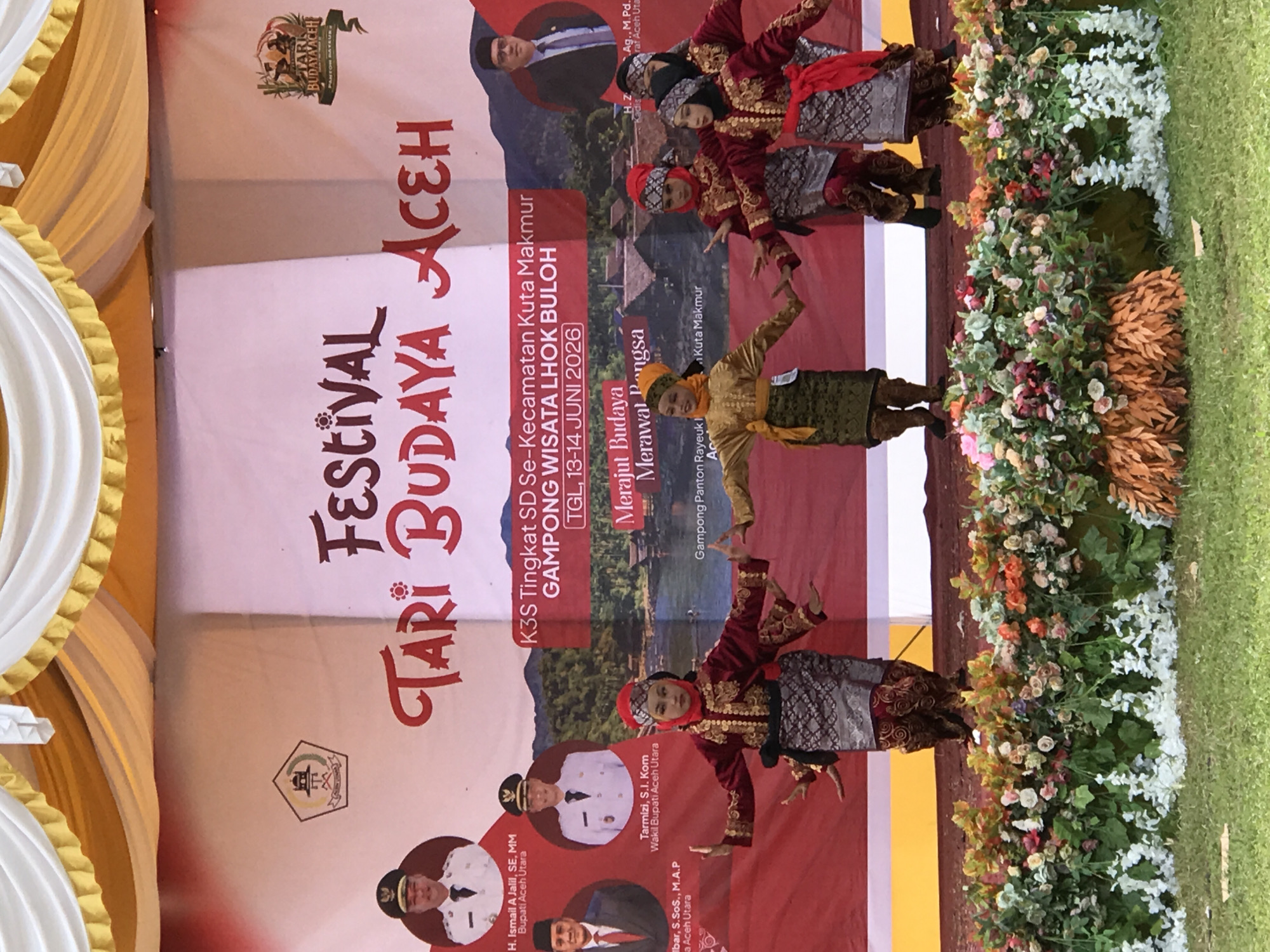 This is a photograph of a cultural festival performance in Lhok Buloh, Kuta Makmur, Aceh, Indonesia. The stage is decorated with a red backdrop featuring a banner with Indonesian text and images of dignitaries. In the foreground, a group of children dressed in traditional Acehnese attire are performing a dance. They are wearing elaborate costumes with intricate embroidery and head coverings. The stage is adorned with a floral arrangement. The overall atmosphere suggests a vibrant cultural event.