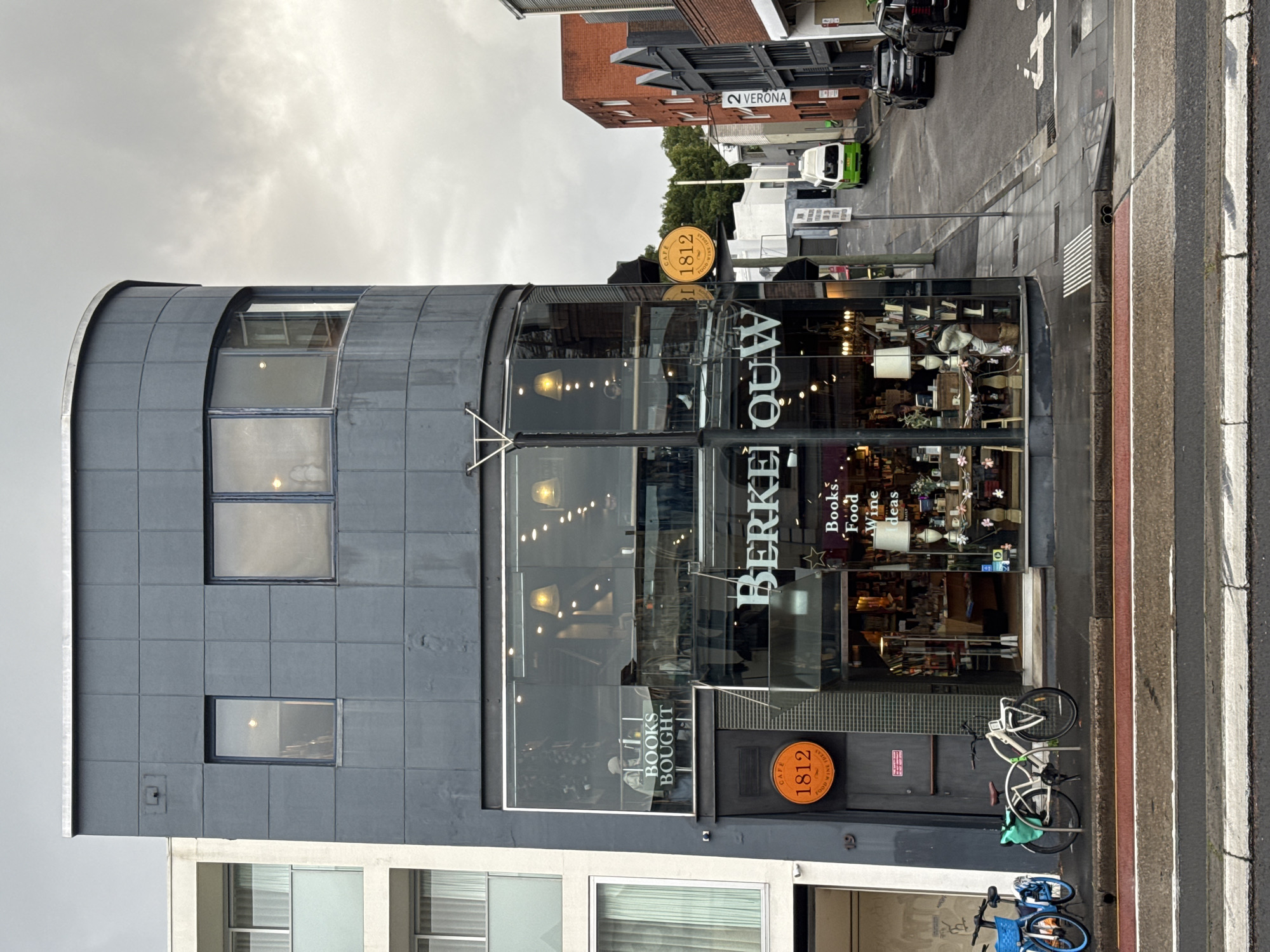 The image depicts the exterior of a multi-story building on a cloudy day, located in Potts Point, Australia. The building features a dark gray, tiled facade on its upper levels and a large glass storefront at street level. The storefront displays "BERKEIJOUW" in large white lettering and a smaller sign that reads "BOOKS. Food Wine Ideas." Inside the store, shelves are filled with books, and various lamps and decorative items are visible.
To the left of the storefront, a circular orange sign with the number "1812" and text reading "CAFE FOOD WINE IDEAS" is mounted on the dark facade. Adjacent to this sign, the number "19" is visible. Two bicycles are parked on the sidewalk in front of the building, one blue and one white with green saddlebags.
The street in front of the building has a pedestrian crossing with red and gray stripes. A sidewalk with gray paving stones runs alongside the storefront, leading to a street that slopes upwards and curves to the right. On the street, a white and green van is parked, and two black cars are parked further up the road. Red brick buildings are visible on the right side of the street. The sky is overcast with gray clouds.