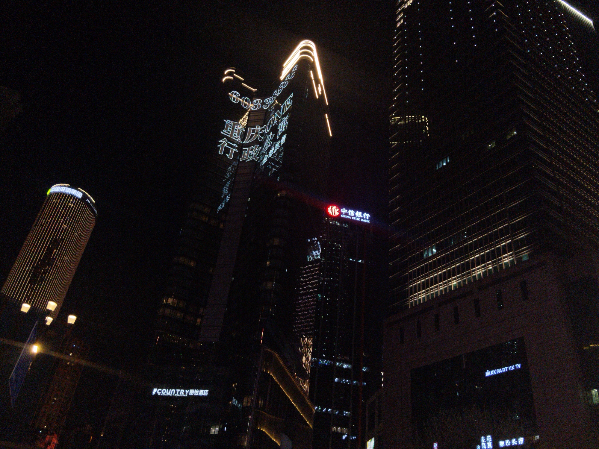 The image shows a night scene in Yuzhong District, China, featuring brightly lit buildings. A cylindrical building on the left is lit with vertical stripes. The central skyscraper displays neon signs in Chinese and numerals including "6033" and "48882", along with what appears to be "ONE". Below it is the logo of China Citic Bank. Further to the right is a taller building partially illuminated with rows of lights forming a grid pattern. At ground level, the words "COUNTRY 丽怡酒店" are visible, while another sign reads "KKK PART Y.K TV". Street lights are scattered throughout the scene.