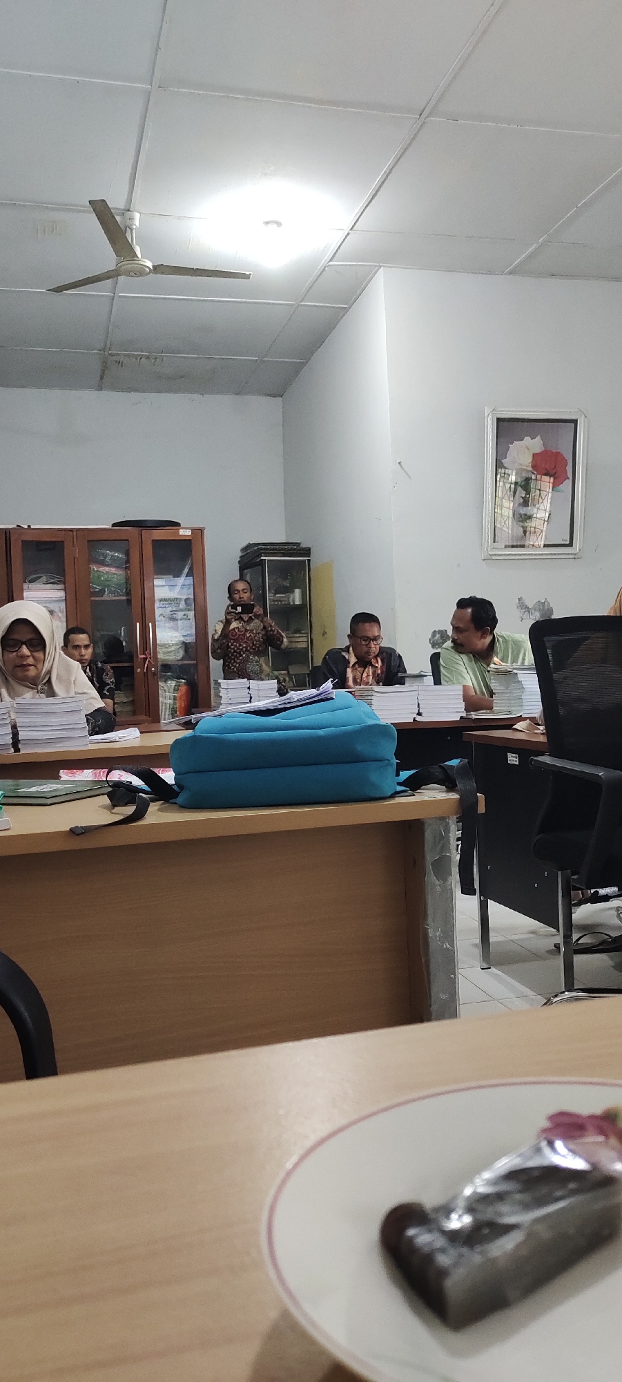 The image depicts an indoor scene, likely an office or meeting room, in Langnibong, Indonesia. A ceiling fan is visible in the upper portion of the frame, with fluorescent lighting illuminating the space. In the mid-ground, several individuals are seated around a table, some appearing to be engaged in a discussion or presentation. One person, wearing a patterned shirt, is standing and holding a smartphone, seemingly taking a photograph or recording. Books and papers are stacked on the table in front of the seated individuals. A blue backpack is placed on the table, and to the right, a black office chair is visible. In the foreground, a partially visible plate with a slice of cake is present on a table. The overall environment suggests a professional or academic setting.