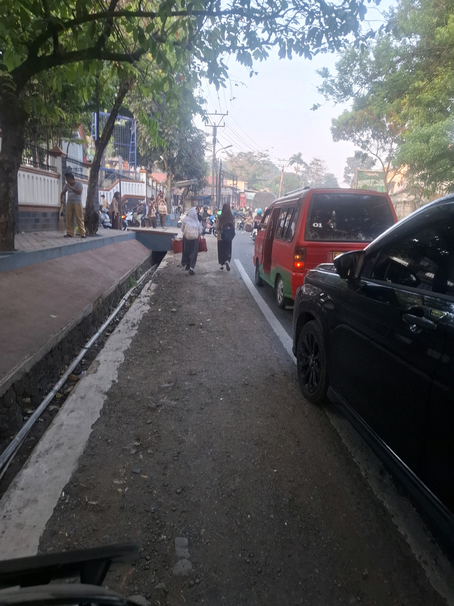 In Sirnagalih, Indonesia, an urban street scene is captured at eye-level. The foreground is dominated by the edge of a dark-colored vehicle, its black wheel and part of its fender visible. To its left, a red and green van is parked, its rear visible with a white "01" sticker. White lines mark the edge of the asphalt road, which is lined with gravel. A concrete gutter runs along the left side of the road, containing a metal pipe. Beyond the gutter, a paved walkway is visible, bordered by a low, white wall. A tree with dark bark and green leaves overhangs the walkway. Several people are present on the walkway and the road. Two individuals, one in a white headscarf and dark clothing, the other in a dark headscarf and black clothing, walk away from the camera on the road. Further back, a man in tan pants and a grey shirt stands on the walkway. Other individuals are seen near parked motorcycles in the middle ground, suggesting a busy area. The background reveals buildings and more trees, with a hazy, bright sky suggesting it is either early morning or late afternoon.