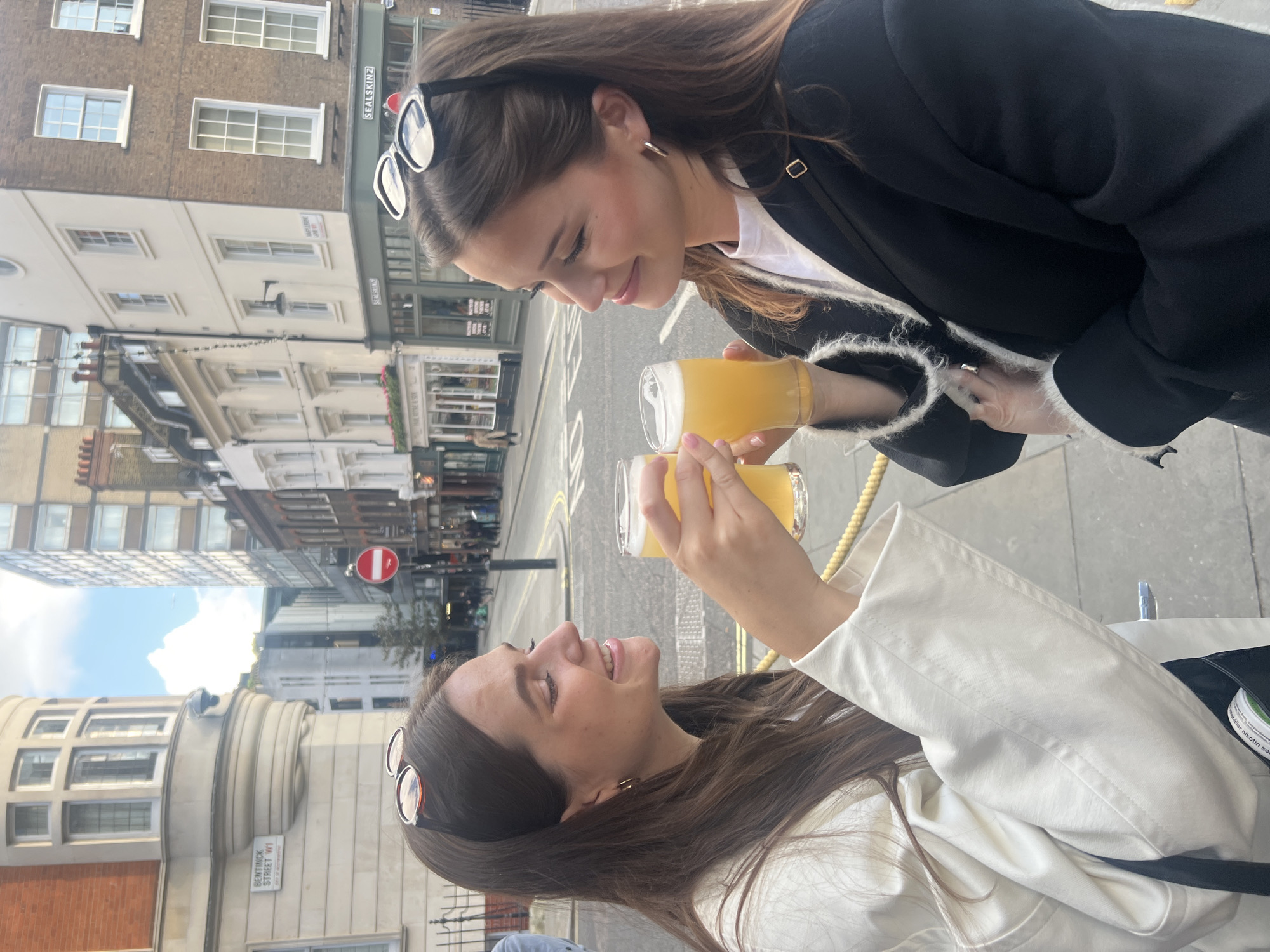 Two young women are toasting glasses of beer in the city of London, United Kingdom. One woman is wearing a black blazer over a white top, and sunglasses are perched on her head. The other woman wears a white jacket, also with sunglasses on her head. The street scene behind them includes buildings, street signs like "Bentinck Street W1", and a red "no entry" sign, helping identify the location as a London street. They are interacting by looking at each other and bringing their beer glasses together for a toast.