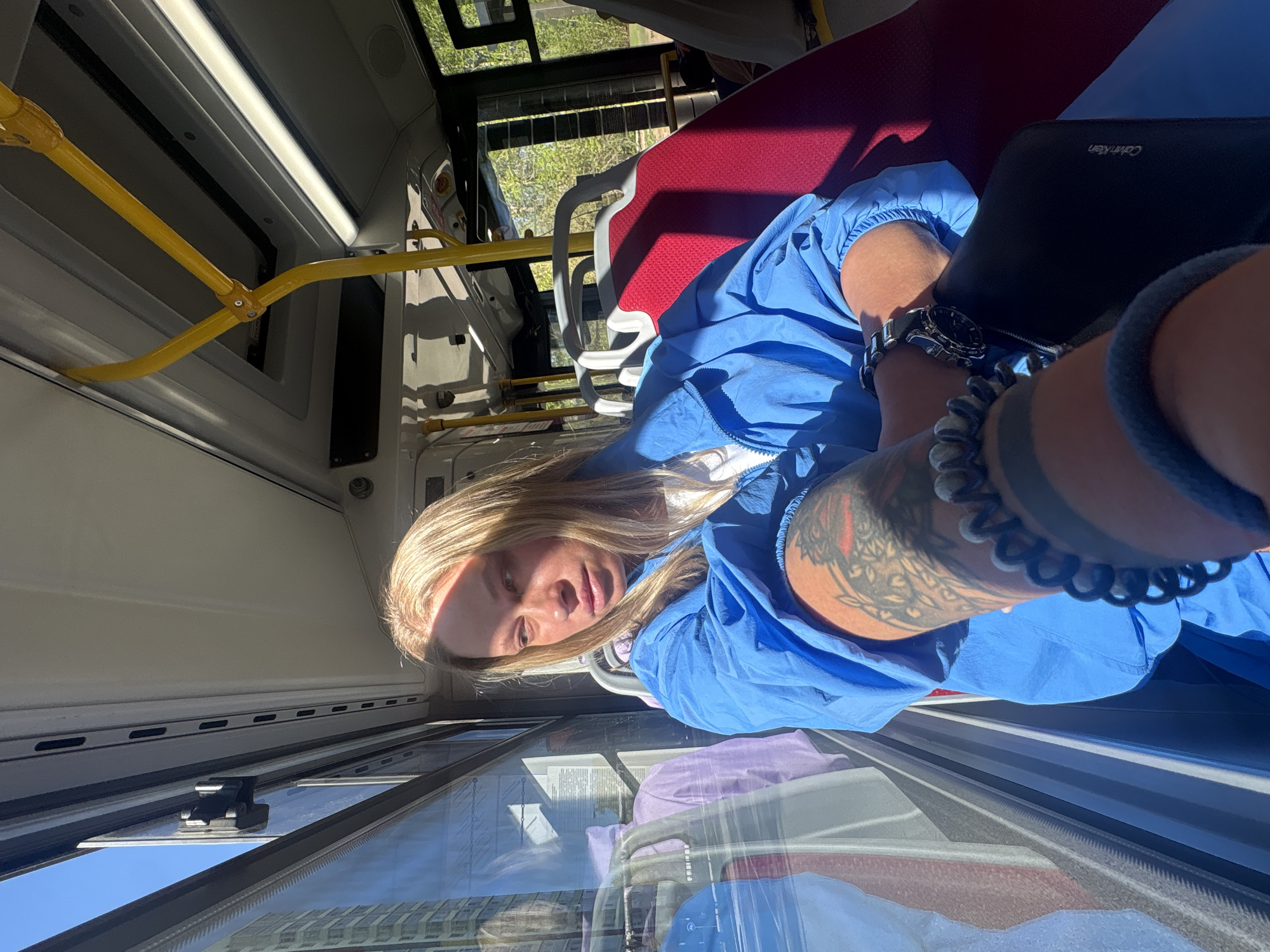 A woman with long blonde hair is sitting in a public transport vehicle. She is wearing a blue top, a wristwatch, and bracelets on her left wrist. A black Calvin Klein wallet rests on her lap. A tattoo is visible on her left arm. The interior of the vehicle has red seats, yellow handrails, and large windows. Outside the windows, buildings are visible, suggesting an urban environment. The reflections in the windows add complexity to the scene. It is not possible to confirm the specific location of the city of Perm, Russia.