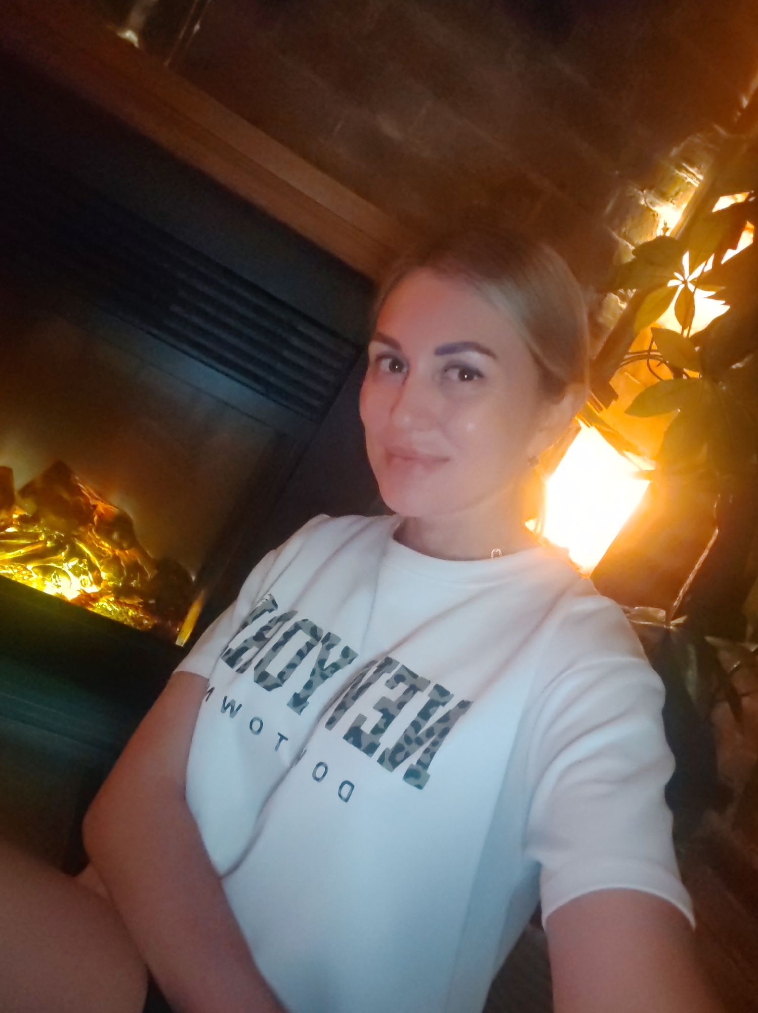 A woman with blonde hair and makeup is posing for a selfie. She is wearing a white t-shirt with a leopard-print pattern and lettering that appears to say "BEYOND". She is sitting in front of a fireplace with artificial flames. To her right, there is a plant with green leaves illuminated by a warm light. The background appears to be a brick wall. The image was taken indoors.