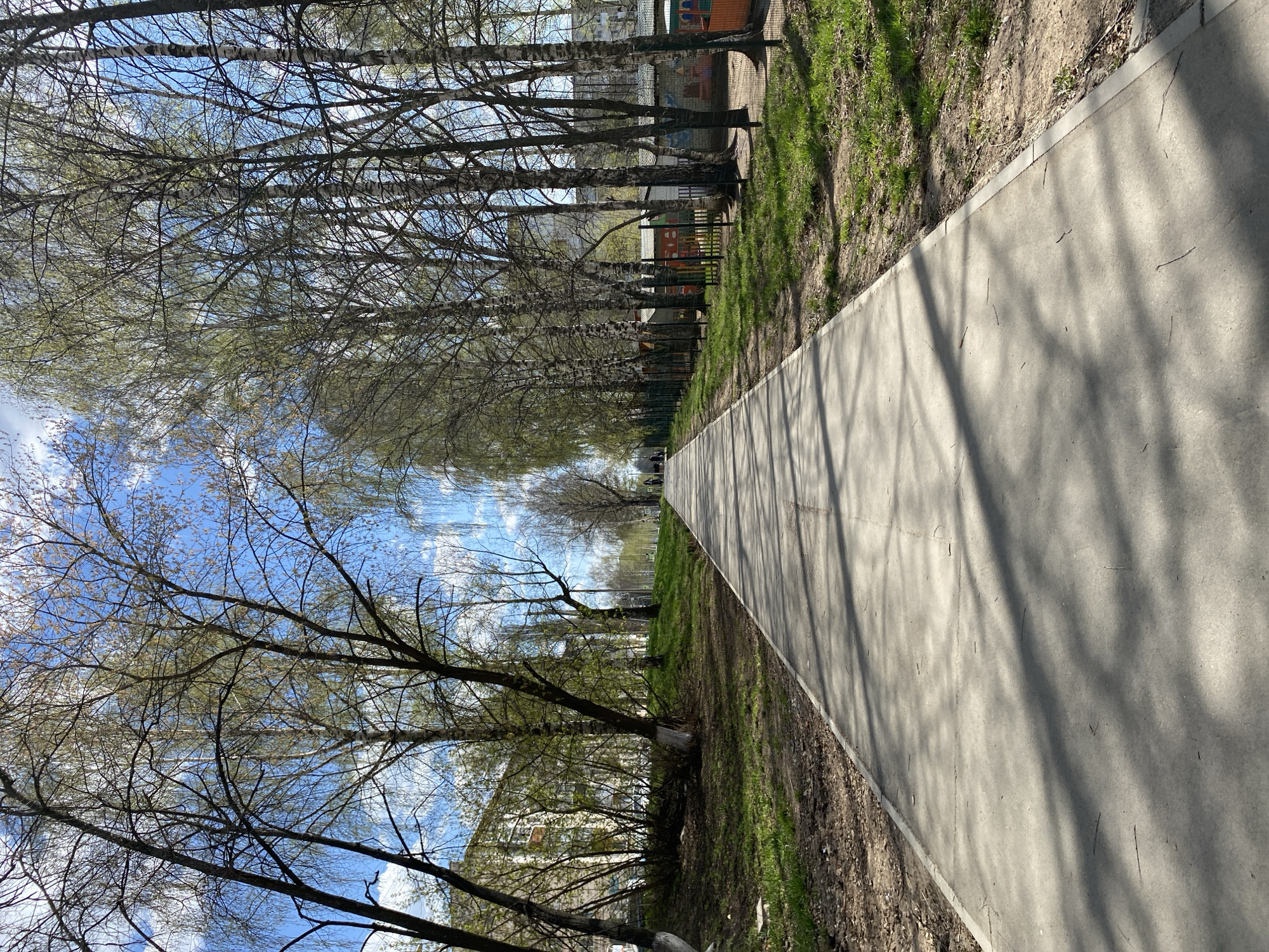 The image shows a concrete pathway running through a park-like area in Votkinsk, Russia. The path is bordered on both sides by grass and trees, some of which appear to be birch trees. A fence runs along one side of the path, behind which is a playground or recreational area with brightly colored structures. The sky is visible through the branches of the trees, showing a mix of blue and white clouds. The image suggests a sunny day, with shadows of the trees cast onto the concrete pathway.