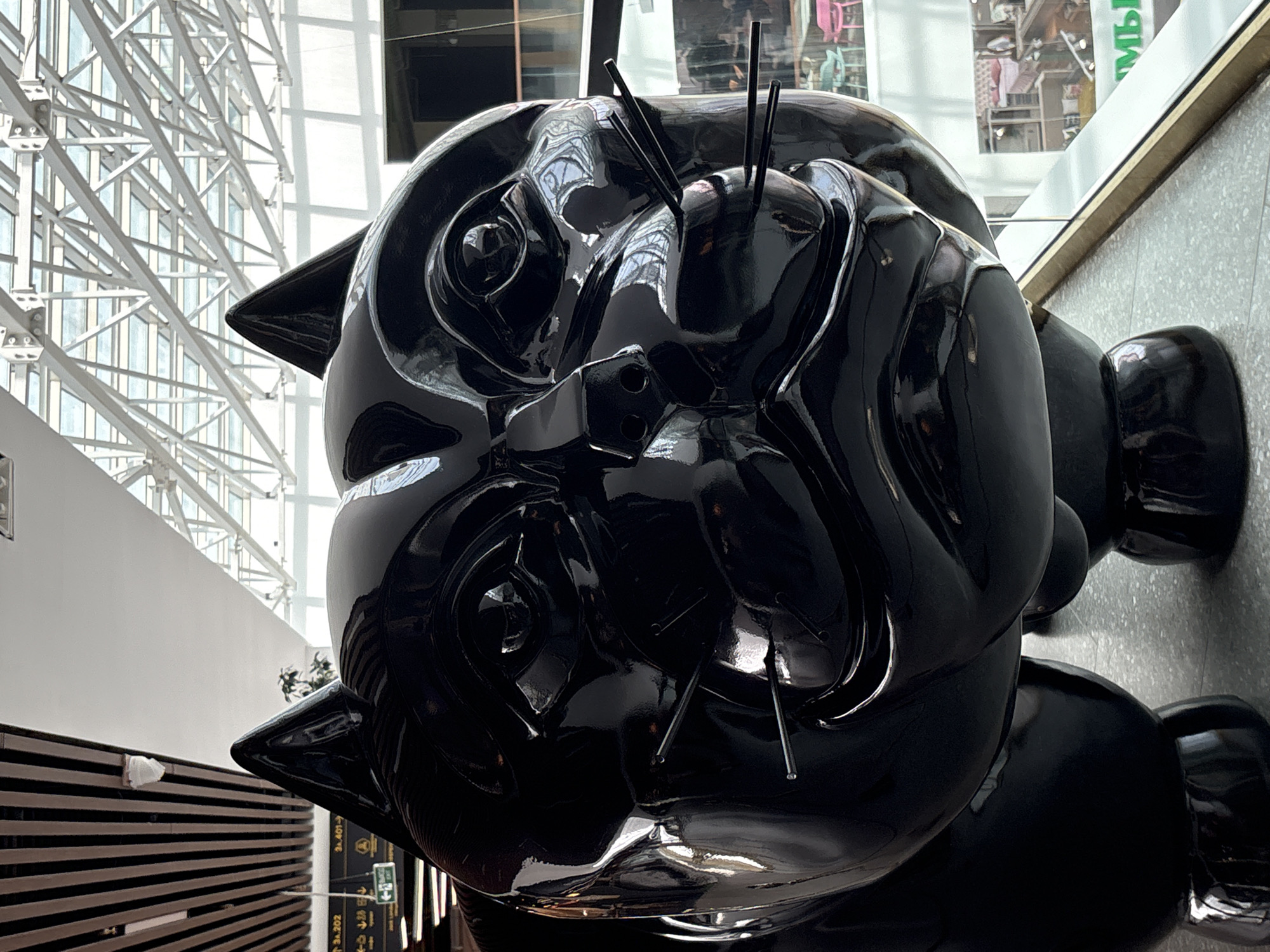 A large, glossy black sculpture of a cat's head is prominently displayed indoors, likely in a shopping mall or public atrium. The sculpture features exaggerated features like large, round eyes and prominent whiskers made of thin rods. The cat's head is turned slightly to the right, with its gaze directed forward. In the background, a modern architectural ceiling with exposed metal beams and large windows is visible, suggesting an open, airy space. Below the cat sculpture, a modern interior with shops and seating areas can be seen through a glass railing. The overall impression is of a contemporary public art installation in an urban setting. The image was taken at Pulkovo Airport in Saint Petersburg, Russia.