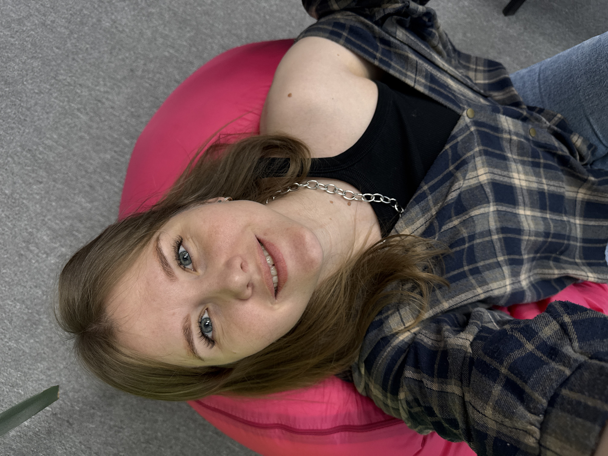 A young adult female is captured in a casual, overhead shot, lying on her back on a vibrant pink bean bag chair in an indoor setting, likely in Perm, Russia. Her head is positioned towards the lower left of the frame, tilted slightly, and she is looking directly at the camera with a relaxed, gentle smile.
The subject has light brown hair, striking blue eyes, and fair skin with visible freckles across her cheeks and nose. She is wearing a black tank top underneath an open, long-sleeved plaid flannel shirt featuring a pattern of blue, beige, and darker blue squares. A silver chain necklace adorns her neck. Her legs, partially clad in blue jeans, extend towards the upper right of the frame. The immediate background consists of a neutral gray carpet or rug, with a small portion of a green plant visible in the bottom left corner. The lighting is even, suggesting an indoor environment. There is no visible text in the image. The overall impression is one of comfort and ease.