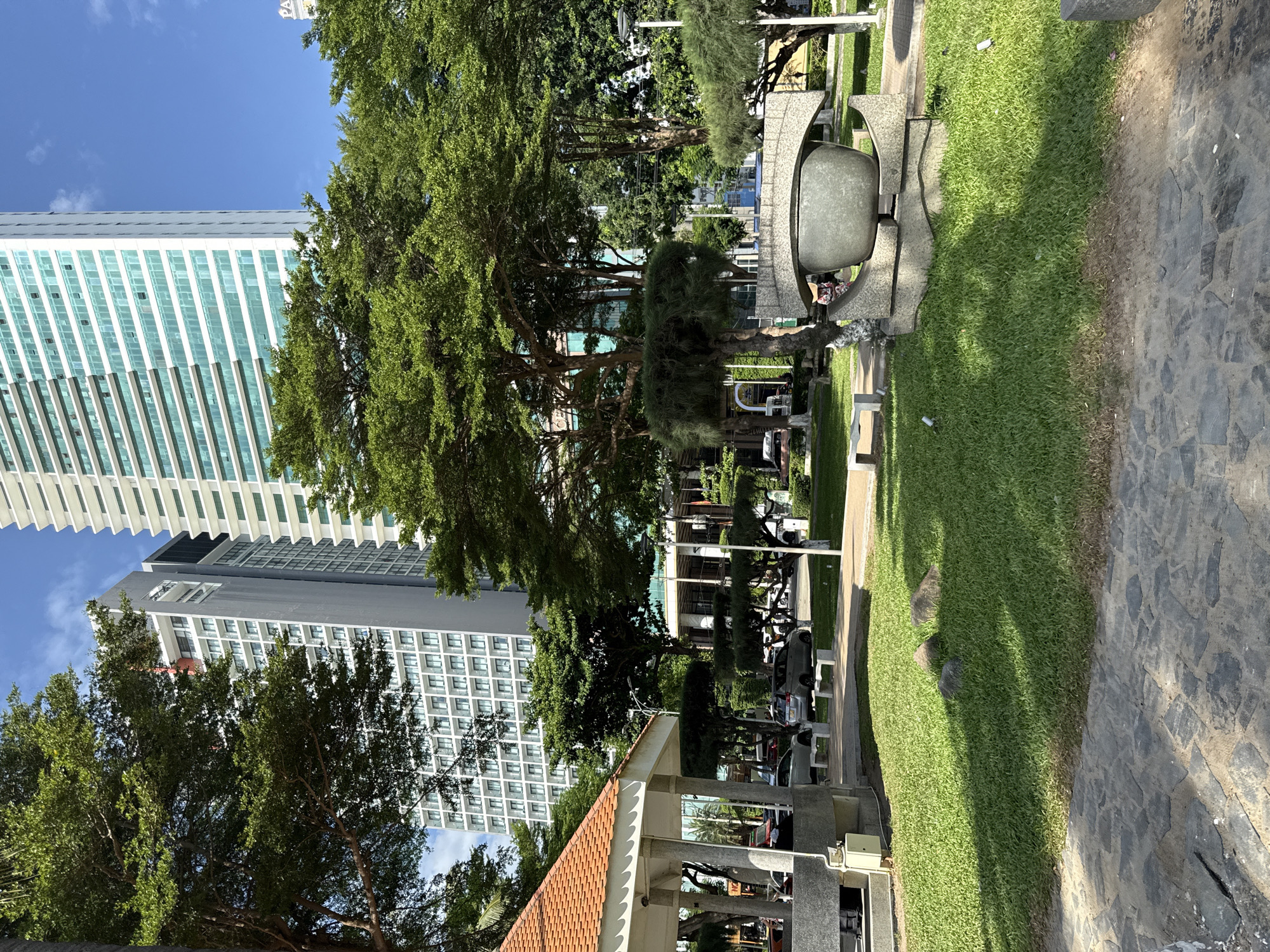 The image depicts a view of Nha Trang's beachfront park area. In the foreground, a stone pathway leads to a manicured green lawn. To the left, a covered pavilion with an orange tiled roof stands on concrete pillars, adjacent to a light gray utility box. Further into the park, a large, modern sculpture with a gray, bowl-like centerpiece is visible, surrounded by lush trees. In the midground, cars are parked along a road, and benches are placed on the grass. Dominating the background are two tall, modern buildings. The one on the left is a gray high-rise with numerous white-framed windows, while the one on the right is a white building with a facade of green-tinted windows and balconies. The sky is clear and blue, suggesting a sunny day.