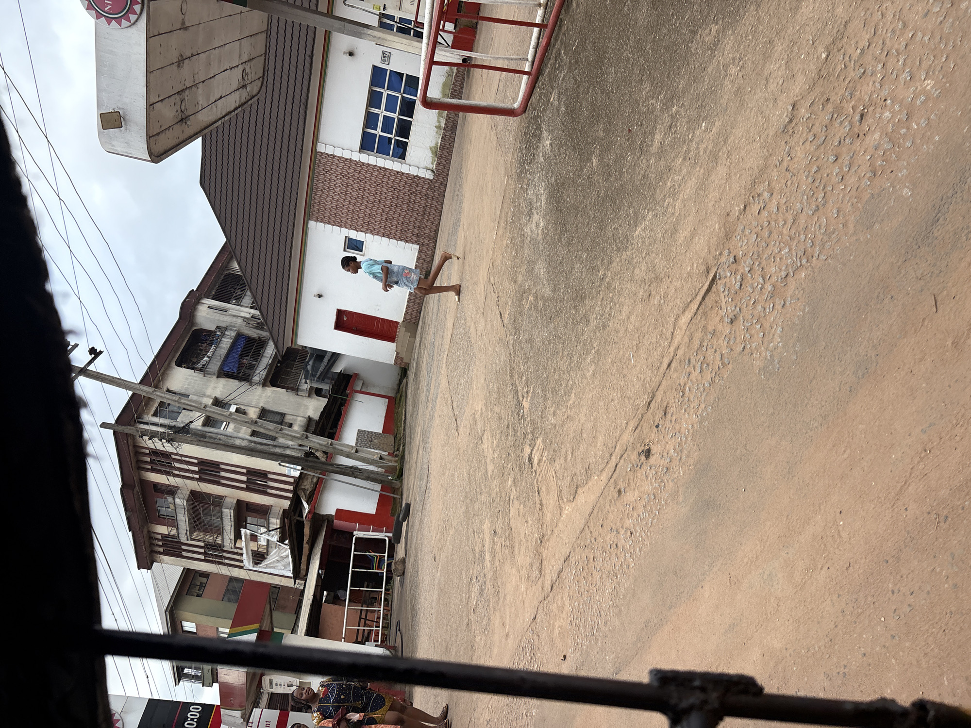 This image captures a street scene in Nkpor, Nigeria. In the foreground, a dirt road with patches of loose gravel and wear is visible. A black metal bar, possibly from a vehicle or barrier, runs vertically along the left edge of the frame, partially obscuring the view.
To the right, a young woman in a light blue t-shirt and patterned shorts is walking away from the camera, her body angled towards the left. She is barefoot and appears to be moving across the road.
Behind her, a white building with a brown tiled roof is situated, featuring a red door and a large window with blue-tinted panes. The building is adorned with decorative brick-like tiling. Further to the right, a portion of a red and white barrier is visible.
In the background, taller buildings with multiple floors and balconies are visible. One building is painted in alternating cream and maroon vertical stripes. Power lines crisscross the sky above the structures. To the far left, the edge of a dark sign with white lettering is partially visible. Two women in colorful clothing are standing near a white structure with red accents. The overall impression is a busy, urban environment.