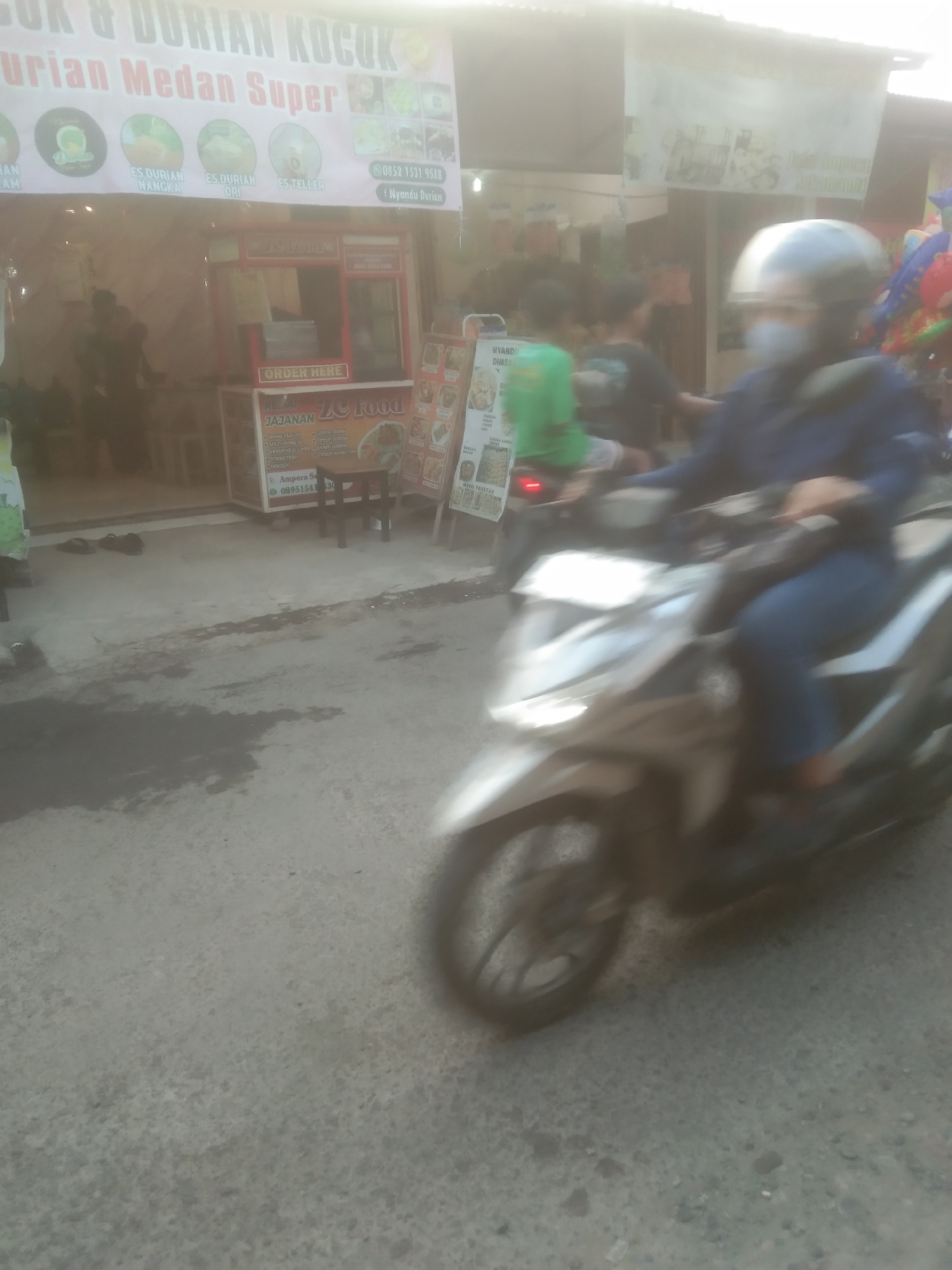 A street scene in Subang, Indonesia, shows a person on a scooter with a helmet and face mask riding on an asphalt road. The scooter appears to be a Honda Beat, white and silver, with a blurred background. To the left of the road is a food stall with a large banner advertising "ES DURIAN & DURIAN KOCAK". The stall has a menu with various durian-based items. In the background, there are other food stalls and shops, as well as people. The lighting suggests it is daytime, possibly late afternoon, with sunlight casting shadows. The overall impression is a bustling street in a commercial area of Subang.