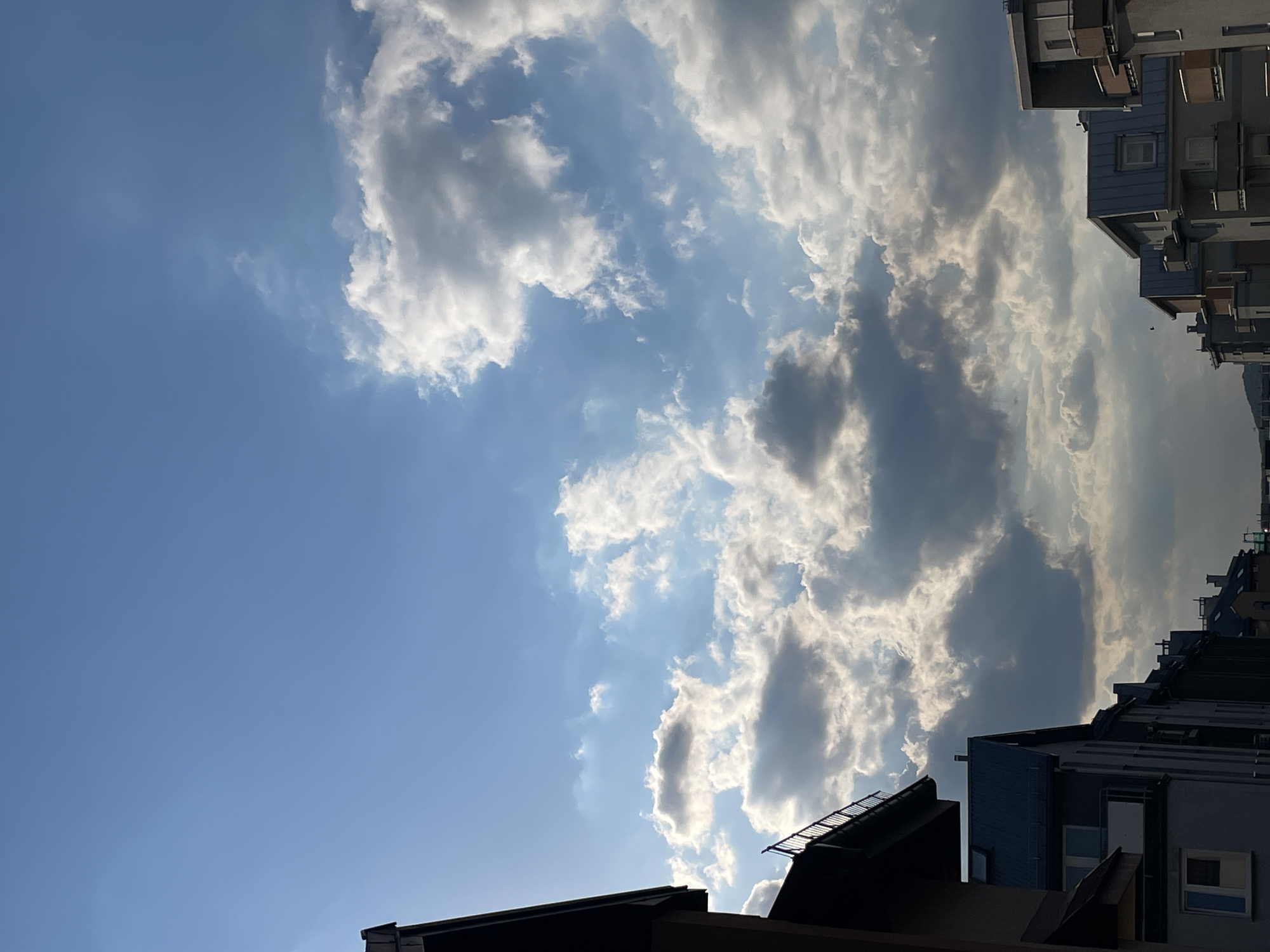 The image shows a view looking up at the sky between buildings in an urban area. The sky is blue with scattered white and gray clouds, some of which are brightly illuminated by sunlight. The buildings on the right and bottom of the frame are multi-story structures with varying rooflines and architectural features.