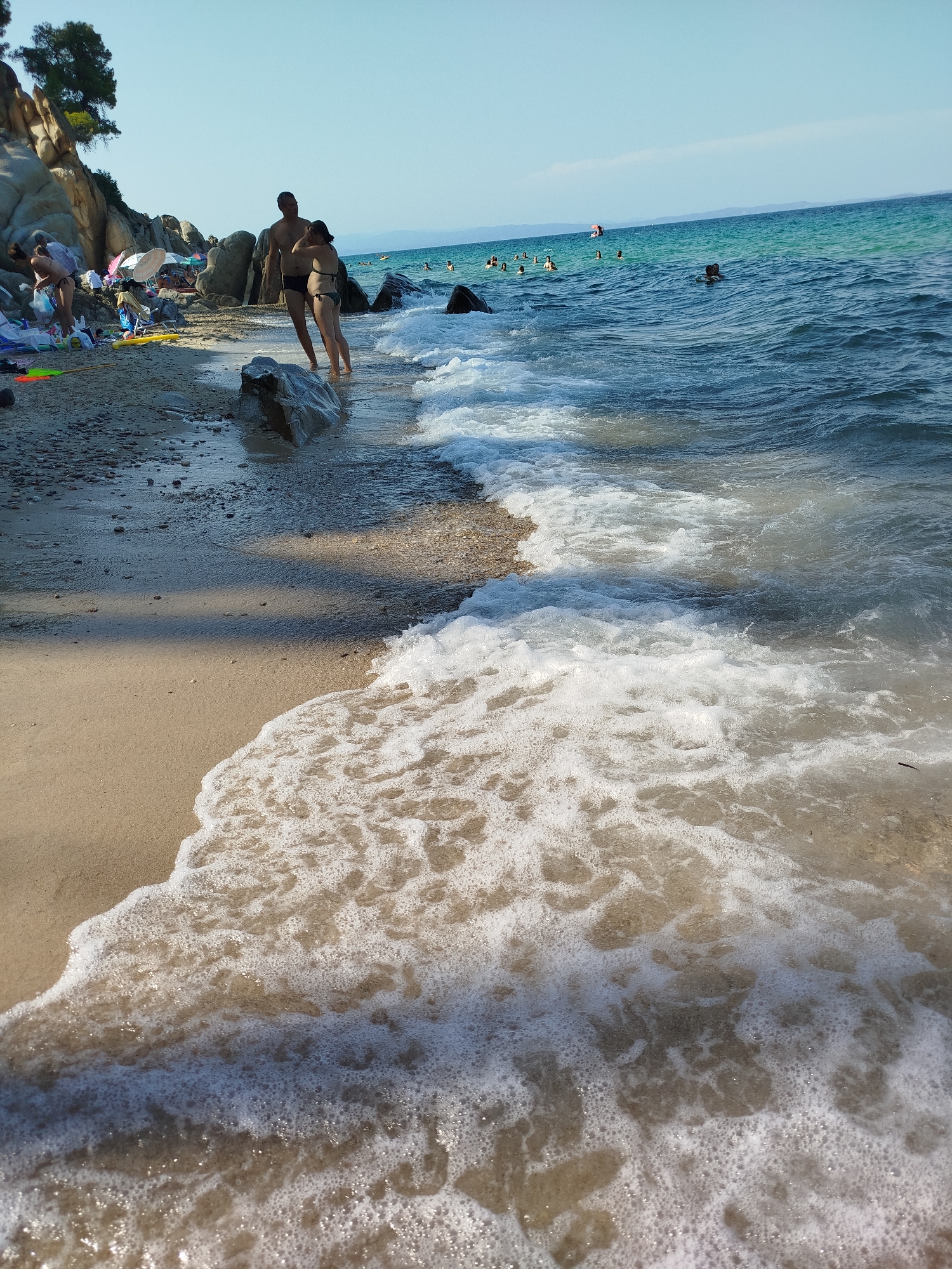 The image depicts a crowded beach scene on a sunny day in Sithonia Municipal Unit, Greece, with the turquoise waters of the Aegean Sea lapping at the shore. In the foreground, a foamy wave washes over the sandy and pebbly beach. A couple stands at the water's edge, facing each other. The male is wearing dark swim trunks and the female is wearing a bikini. Further up the beach, several people are visible, some sitting under umbrellas, others walking or standing. Rocks and vegetation dot the coastline. In the background, numerous people are swimming and wading in the sea, and a distant landmass is visible on the horizon.