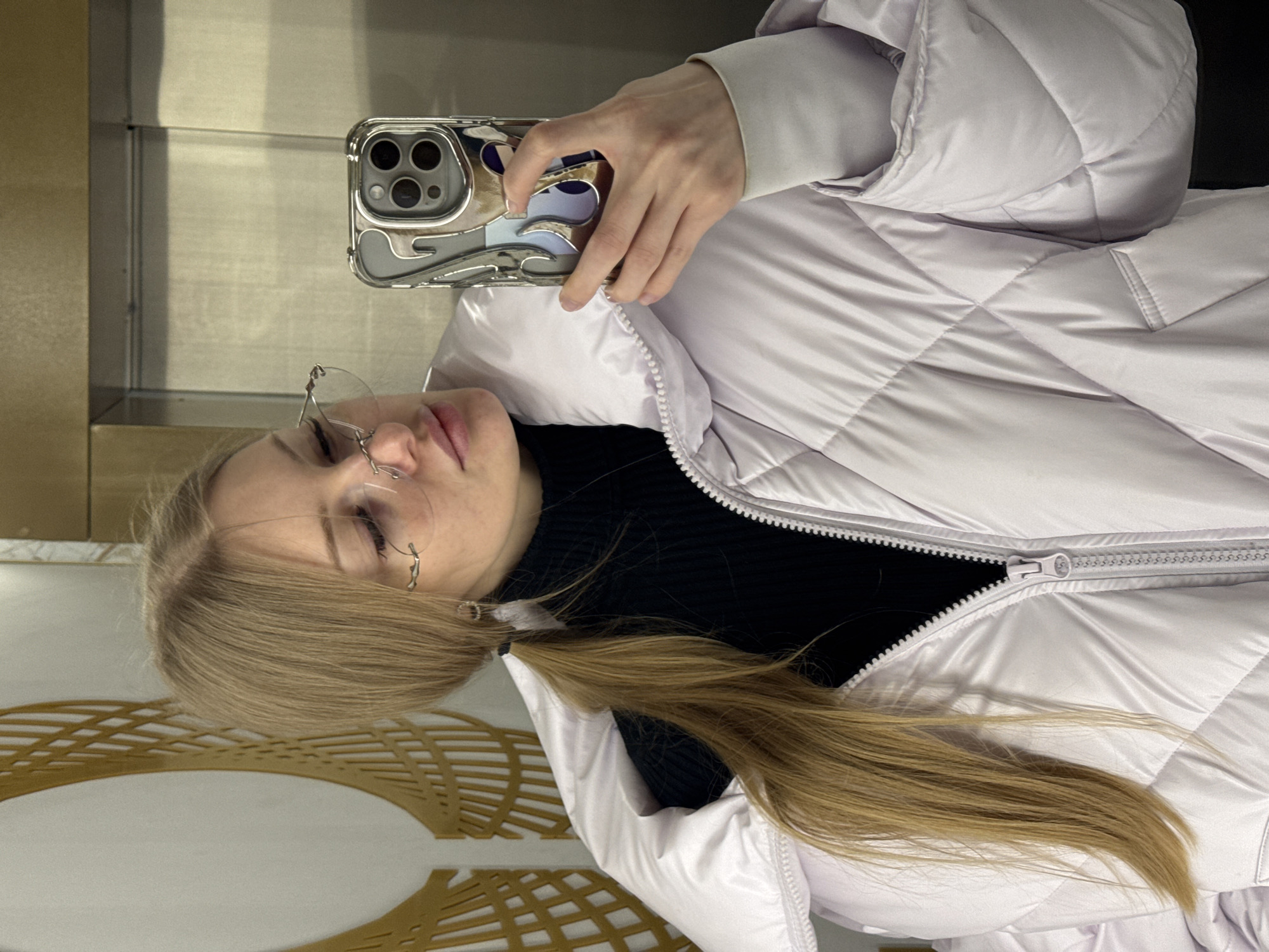 A blonde woman wearing glasses and a lilac puffer jacket takes a selfie in a mirror. She is holding a smartphone in her right hand. The background features a patterned wall and an elevator. The location is within the city of Perm, Russia.