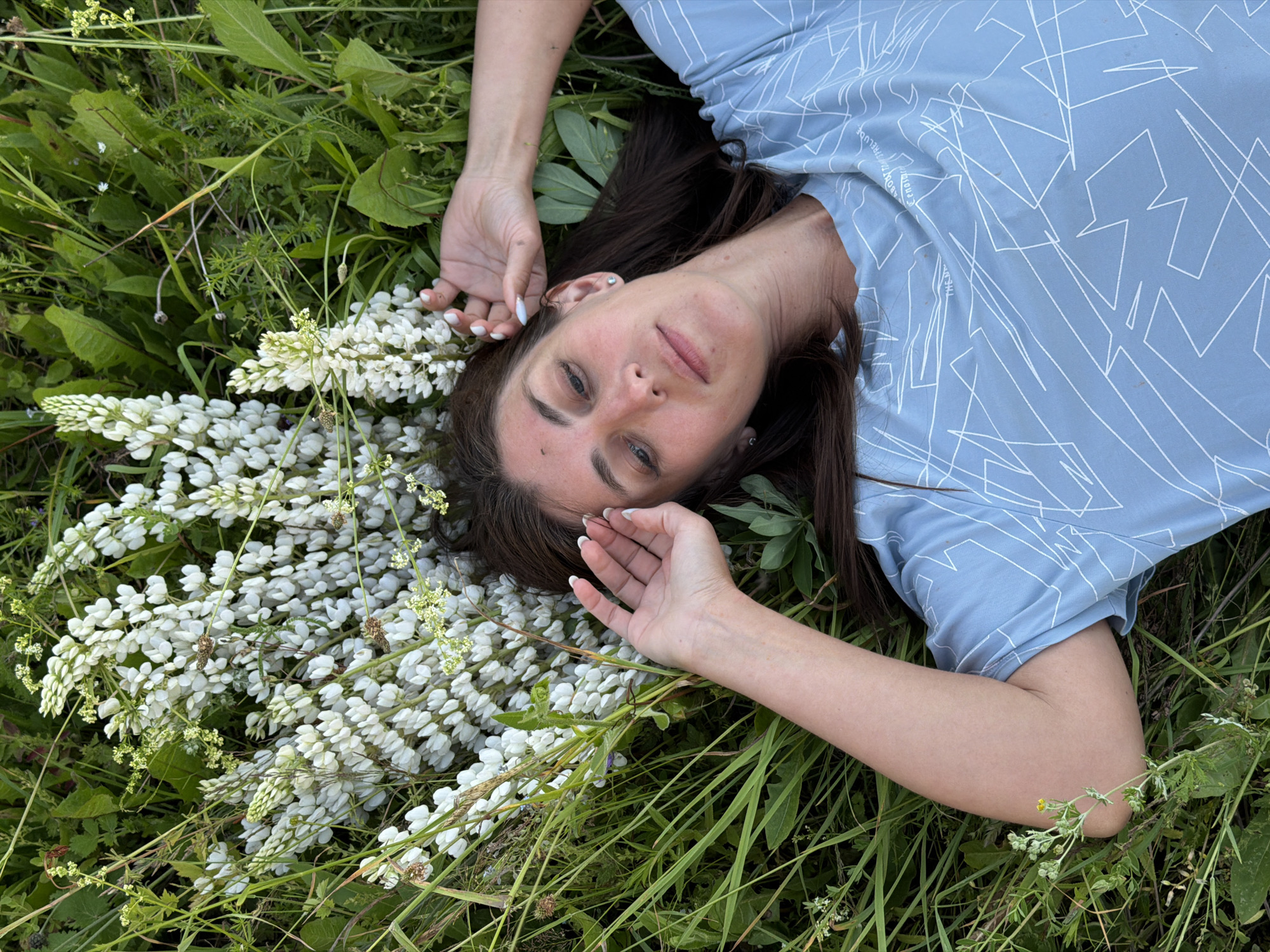 A woman is lying on her back in a grassy field, her head resting on a cluster of white lupine flowers. She is wearing a light blue t-shirt with a white geometric pattern. Her right arm is bent and her hand is resting near her head, fingers splayed. Her left arm is not fully visible. The field is covered in lush green grass and other wildflowers. The woman's eyes are closed, and her expression is serene. The overall mood of the image is peaceful and natural.